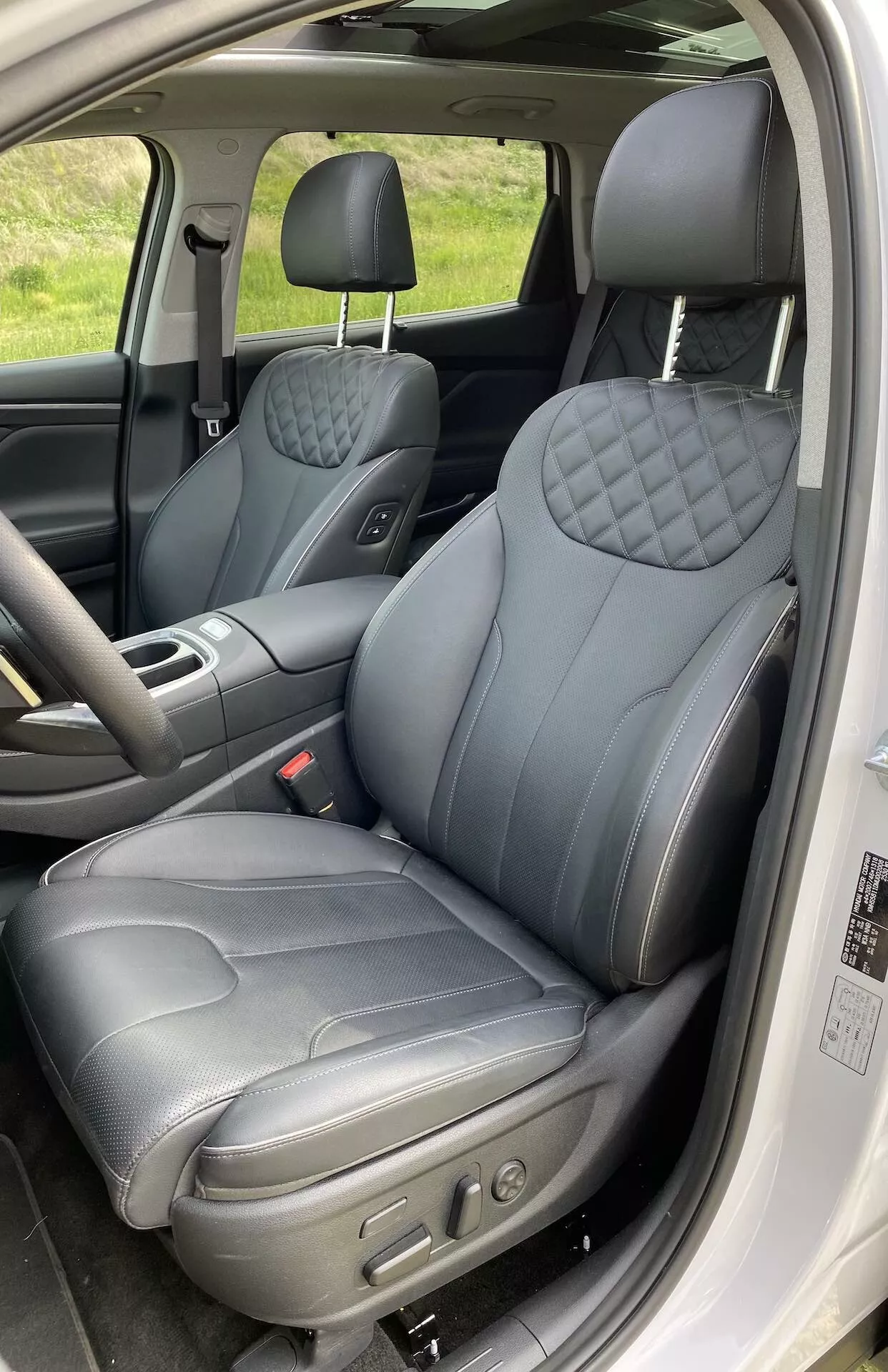 Interior del Hyundai Santa Fe 2020 que muestra el asiento del conductor desde una puerta abierta. La toma vertical en primer plano captura los asientos delanteros, enfocándose en el asiento del conductor que cuenta con tapicería de cuero oscuro con un patrón de diamante acolchado en el respaldo y el cojín del asiento, sugiriendo una variante premium. Los controles electrónicos visibles indican posiciones de asiento ajustables. El reposacabezas parece ajustable, y el panel de la puerta tiene controles de ventanas y posiblemente de espejos. El interior está detallado con costuras contrastantes, realzando la sensación de lujo.