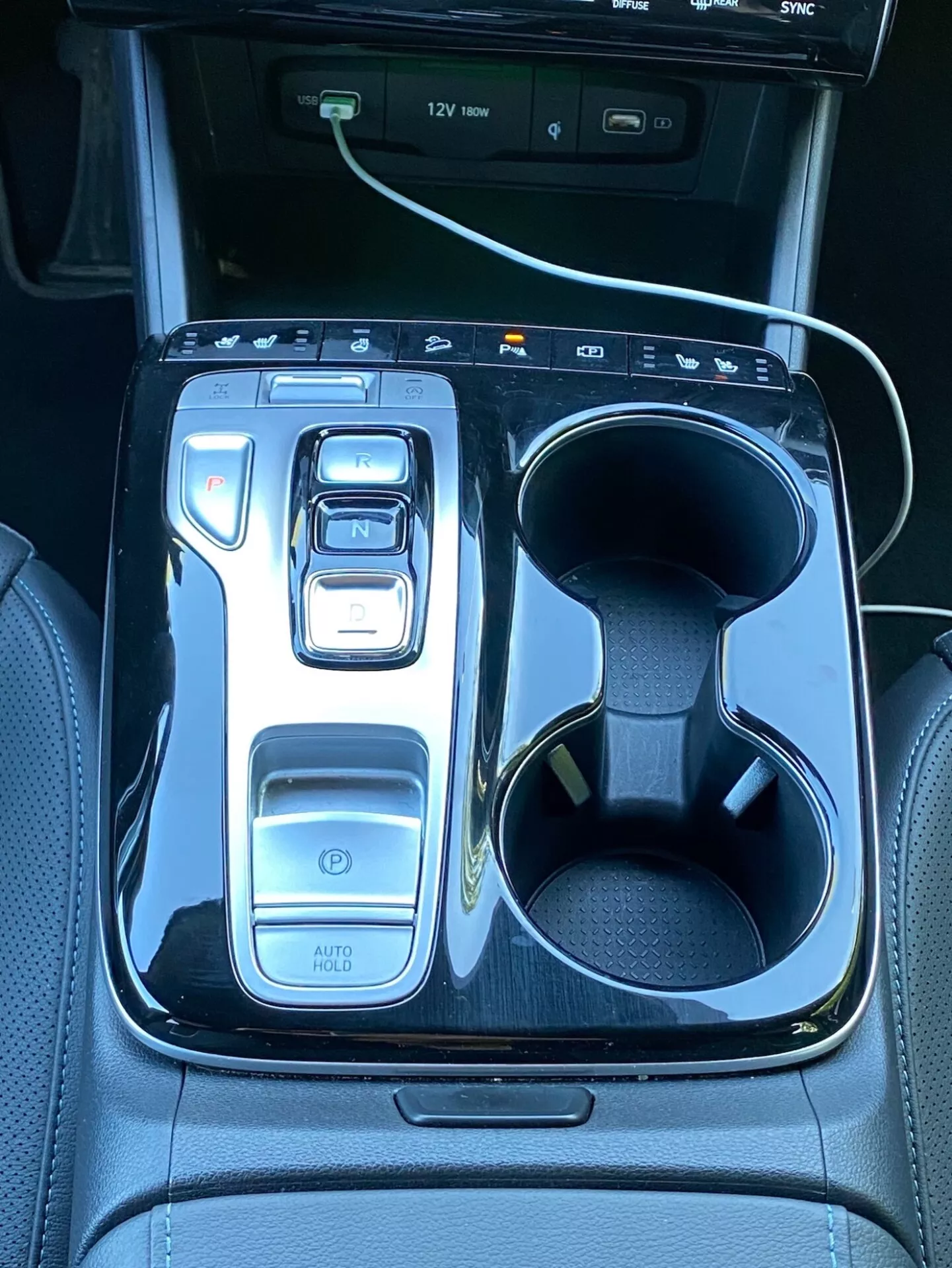 La imagen muestra la consola central de un Hyundai Tucson 2020, con un selector de cambios electrónico en 'N' rodeado por un acabado negro brillante. A la izquierda están los controles que incluyen un botón de encendido, selectores de modos de conducción y la función 'AUTO HOLD'. A la derecha, hay doble portavasos con un cable de carga blanco en uno de ellos. En la parte superior se ve parte de una pantalla de infoentretenimiento con indicadores de puerto USB, destacando la tecnología y el lujo del vehículo.