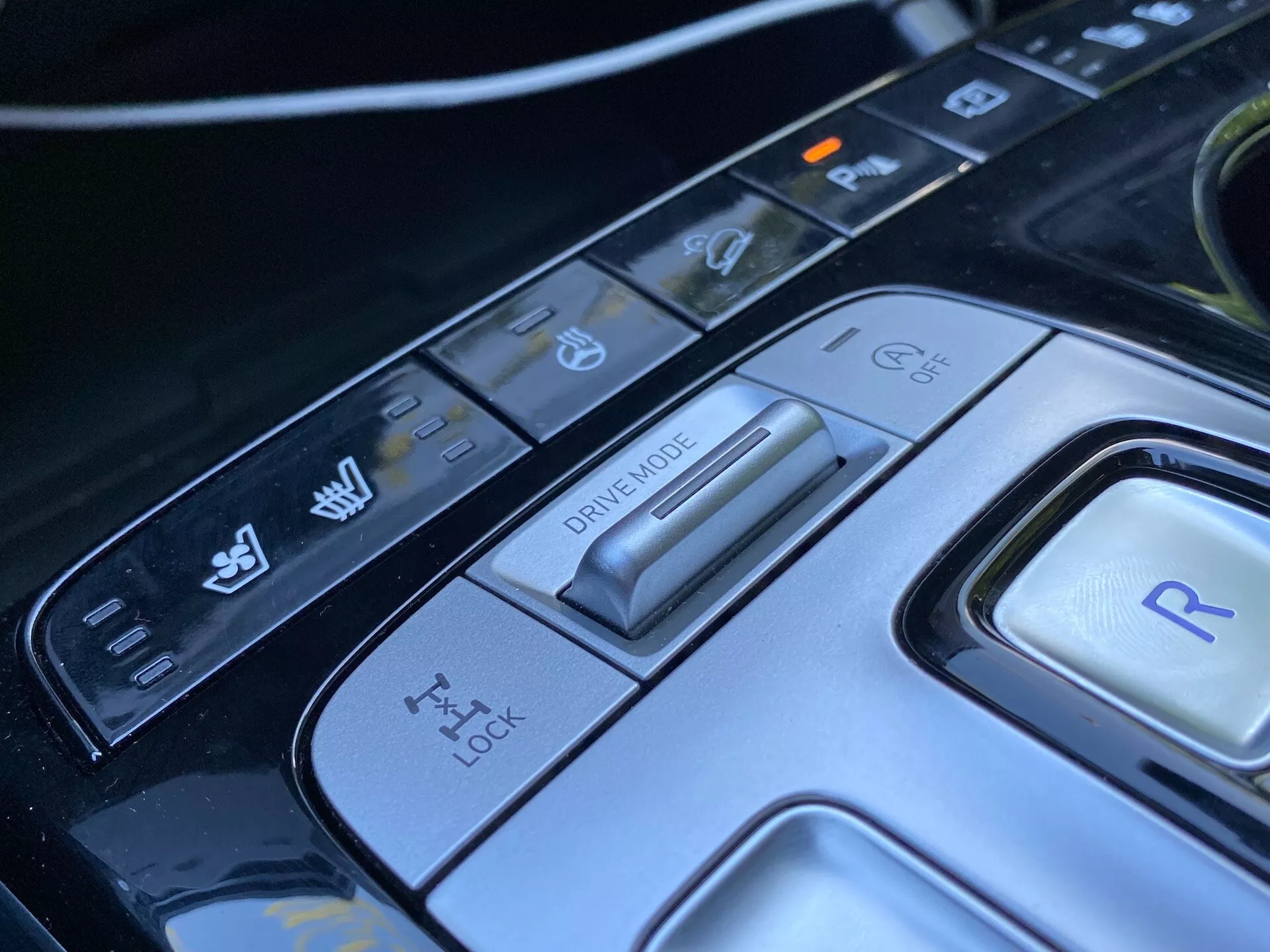 Vista de cerca del panel de control central del Hyundai Tucson 2020, centrada en el selector de modo de conducción y varios botones de control. La imagen muestra un avanzado interruptor de modo de conducción para diferentes condiciones de manejo, un panel de control de tracción, un botón para la marcha atrás y controles adicionales para la estabilidad del vehículo y características de asistencia de aparcamiento. El diseño elegante y los materiales de alta tecnología enfatizan el interior automotriz sofisticado de este coche moderno de lujo.
