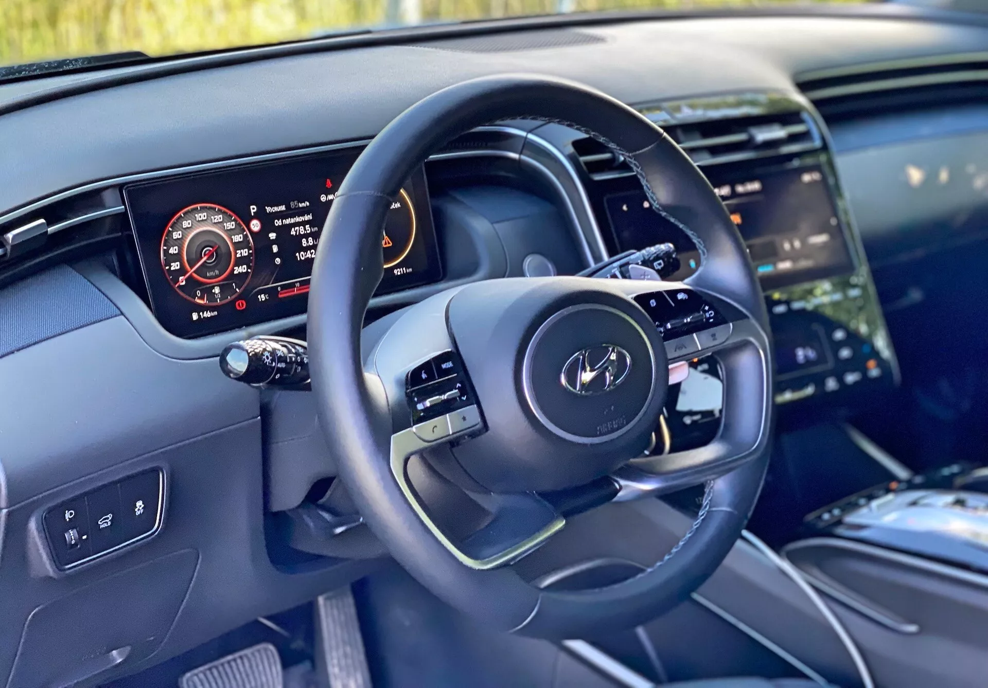 Vista interior de un Hyundai Tucson 2020 desde la perspectiva del conductor que muestra el volante moderno con controles multifuncionales y un panel de instrumentos digital que muestra el velocímetro, el tacómetro, el nivel de combustible y la temperatura del motor. La imagen también resalta una parte de la consola central con un gran sistema de infoentretenimiento táctil, un diseño limpio del tablero con iluminación ambiental y materiales de alta calidad que sugieren acabados de lujo.