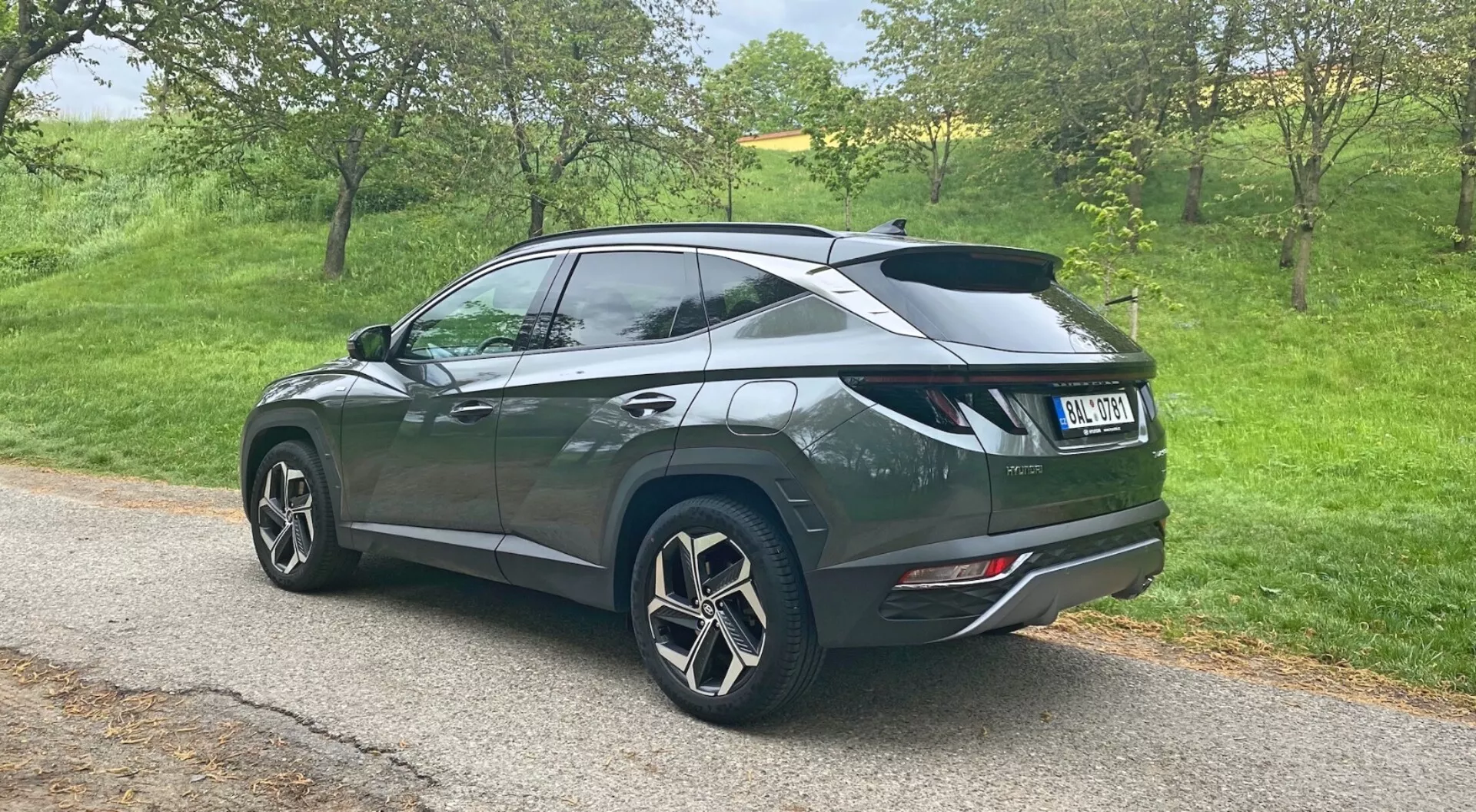 Perspectiva trasera de tres cuartos de un Hyundai Tucson 1.6 T-GDi Pure SUV 2020 estacionado en un camino estrecho en un entorno rural, exhibiendo grandes llantas de aleación de múltiples radios, pintura metálica oscura con una línea de techo descendente, spoiler trasero, vidrios polarizados en las ventanas traseras, luces traseras modernas y puntas de escape dobles integradas en el parachoques trasero.
