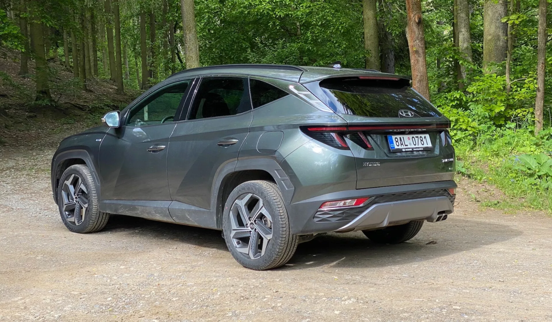 Un SUV moderno estacionado en un camino sin pavimentar en un área forestal, capturado desde una vista trasera de tres cuartos en una ligera elevación. El vehículo presenta un diseño elegante y contemporáneo con líneas afiladas, luces traseras alargadas que envuelven desde la parte trasera hacia los lados, un spoiler visible y reflectores en el parachoques trasero, lo que sugiere características de seguridad avanzadas. El vehículo de color gris metálico tiene ruedas de aleación de múltiples radios y neumáticos de carretera, con una superficie brillante que indica que es nuevo o está bien mantenido. Alrededor del coche hay un bosque verde y exuberante con tierra compactada y grava suelta en el suelo.