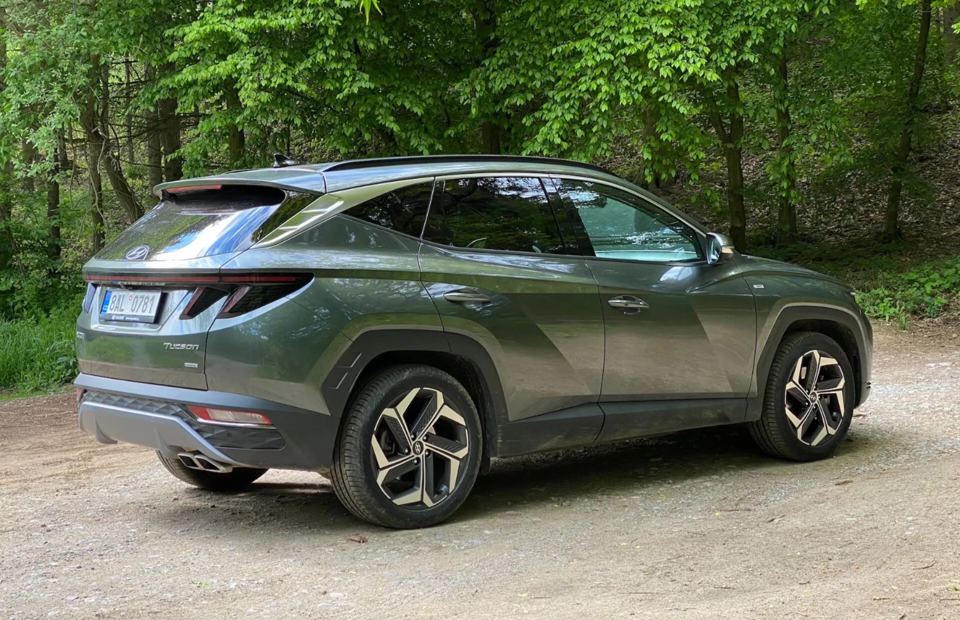 Vista de tres cuartos trasera de un moderno SUV Hyundai Tucson 2020 con enfoque en su perfil lateral y diseño trasero. El vehículo está posicionado a la izquierda con la foto tomada desde la derecha, enfatizando la esquina trasera derecha. Las luces traseras distintivas de LED se envuelven desde la parte trasera hacia los lados y un alerón montado en el techo agrega un toque deportivo. La línea ascendente de la ventana, el techo inclinado, las ventanas tintadas y las manijas de las puertas del color de la carrocería acentúan su apariencia de coupé. Las grandes llantas de aleación con diseño de múltiples radios y los neumáticos de perfil bajo mejoran su sensación de lujo. Detalles menores como el limpiaparabrisas trasero y los moldes de los arcos de las ruedas significan practicidad y robustez. El coche está colocado sobre grava en un entorno natural con árboles en el fondo.