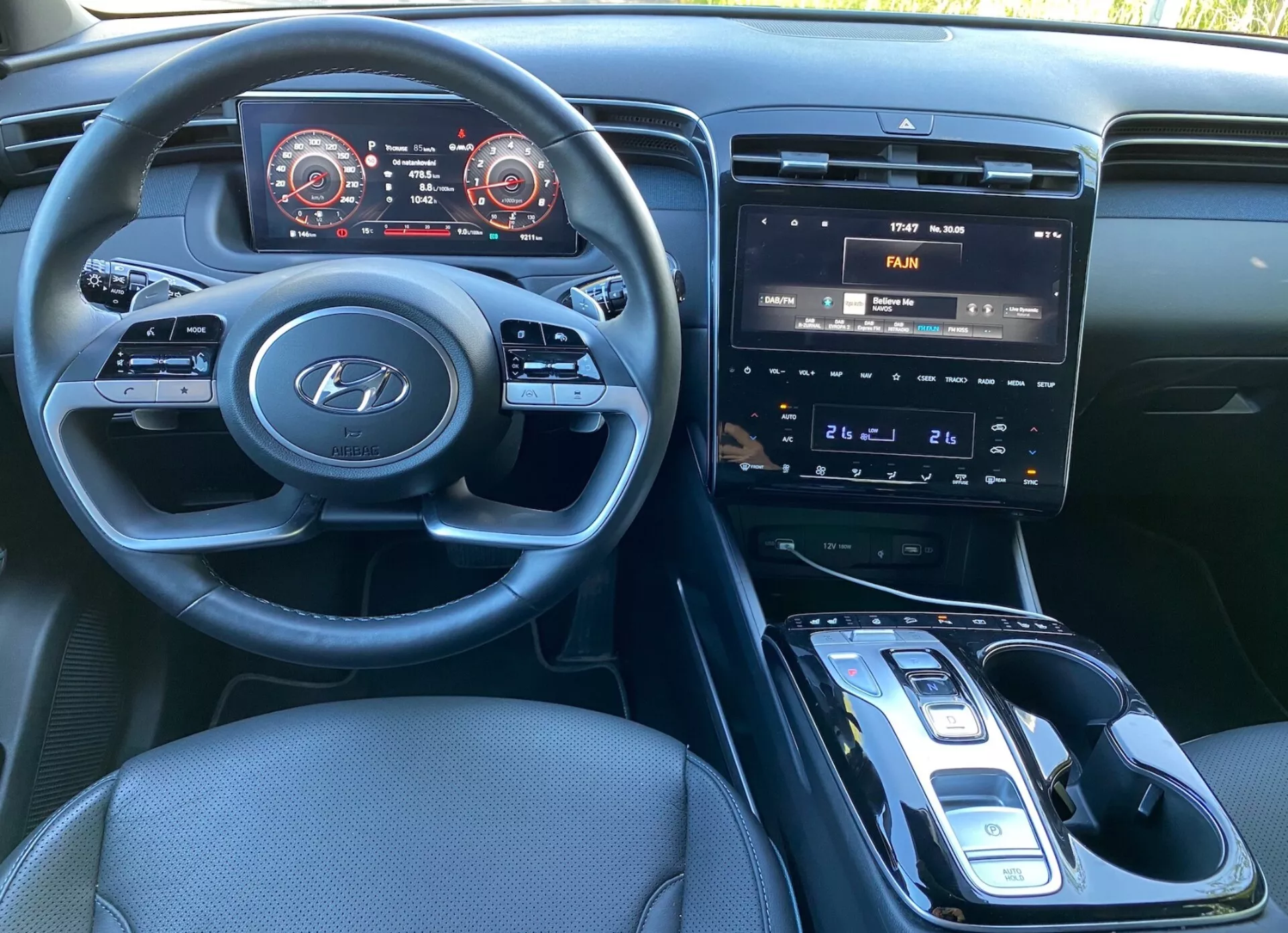 Vista interior del Hyundai Tucson 2020 desde el asiento del conductor, mostrando el volante con el logo de Hyundai y controles para audio, teléfono y configuración del control de crucero. El salpicadero cuenta con tablero de instrumentos digital con velocímetro y tacómetro acentuados en rojo. La consola central tiene un sistema de infoentretenimiento con pantalla táctil que muestra los controles de aire acondicionado, botones físicos y diales para controlar el clima, y una palanca de cambios automática con selector electrónico. Es visible una posible base de carga inalámbrica, tapicería de tela oscura y una cabina bien iluminada.