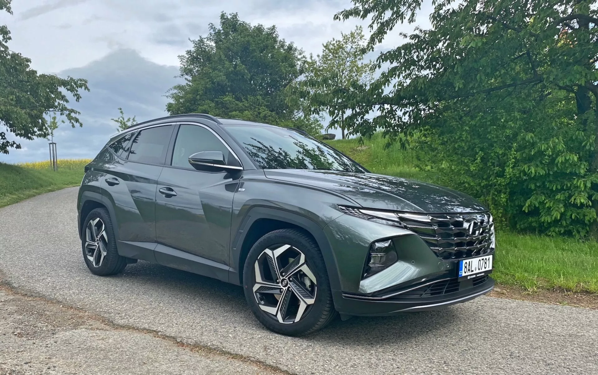 La imagen muestra un moderno SUV Hyundai Tucson 1.6 T-GDi Pure estacionado en una carretera de asfalto con un fondo natural. Fotografiado desde un ángulo de tres cuartos frontal, se pueden ver la parrilla prominente, los faros elegantes, el parachoques delantero con luces antiniebla o tomas de aire, las grandes llantas de aleación con diseño de múltiples radios y los espejos laterales. La silueta del SUV se inclina hacia la parte trasera, indicando un diseño deportivo y aerodinámico. Captada en condiciones de luz diurna suave, la foto resalta el color y los detalles del vehículo sin sombras duras.