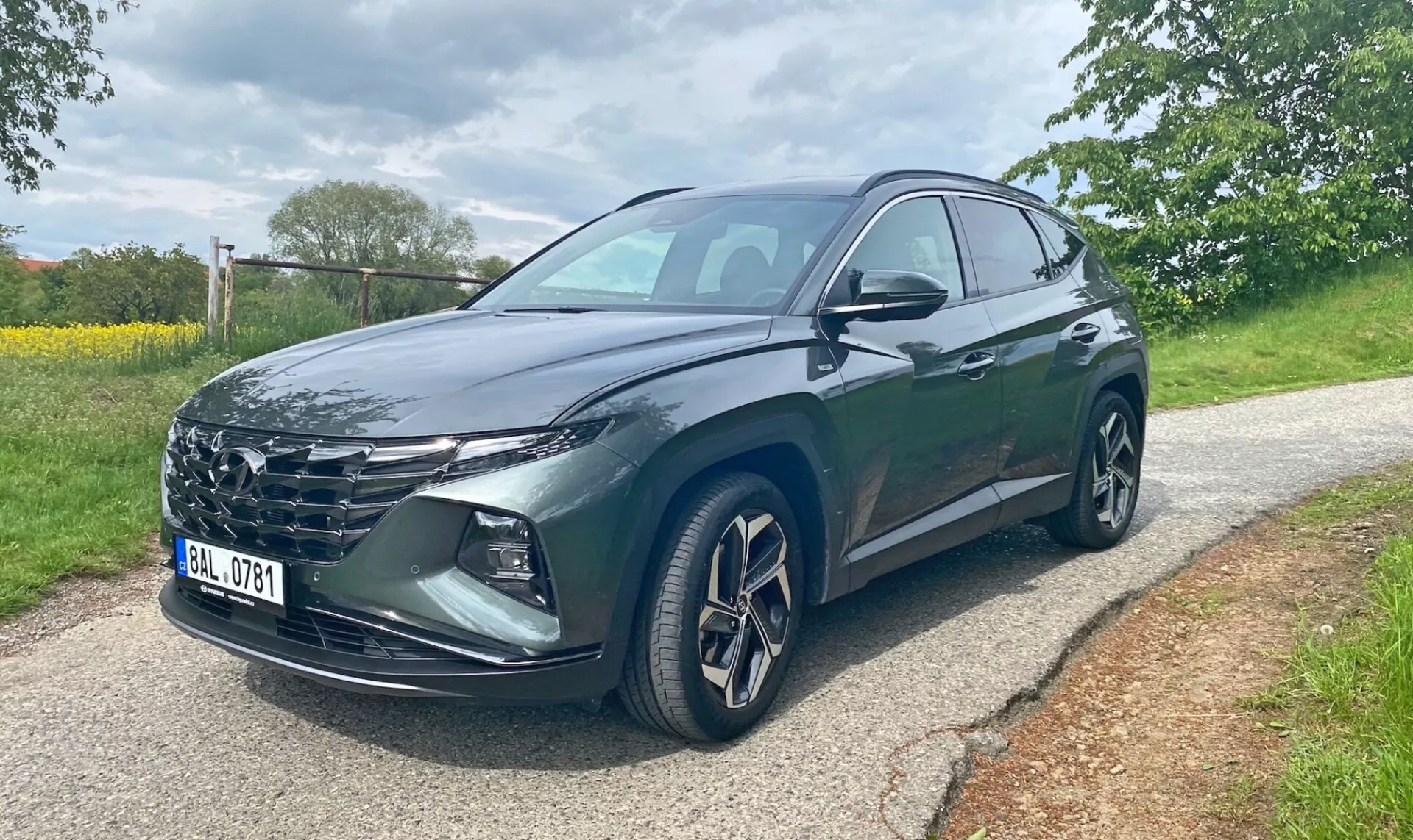 Hyundai Tucson 2020 1.6 T-GDI Pure SUV, visto desde un ángulo de tres cuartos frontales en una carretera pavimentada con árboles y césped en el fondo. La imagen muestra la parrilla del vehículo, los faros LED afilados, las llantas de aleación, los espejos laterales con señales de giro integradas y una pintura metálica oscura. Se ven las puertas delantera del conductor y trasera. Foto tomada durante el día bajo condiciones de iluminación nublada.