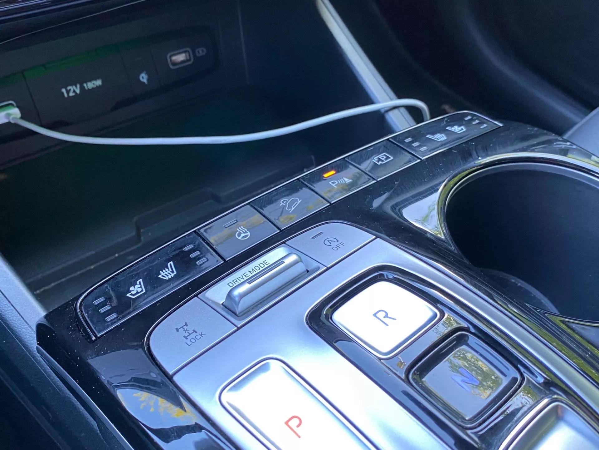 Primer plano de la consola central y área de selección de marchas de un Hyundai Tucson 2020 1.6 T-GDi Pure, que muestra un diseño moderno con botones para parqueo, reversa, neutral, manejo y modos de freno/regenerativo, junto con botones adicionales para apagado automático, sensores de estacionamiento/estacionamiento automatizado y advertencia de mantenimiento/salida de carril. Pantalla de sistema multimedia visible, portavasos y toma de corriente de 12V que indican comodidad, conveniencia y sistemas avanzados de asistencia al conductor (ADAS). La consola tiene un diseño elegante con material negro brillante y acentos metálicos, insinuando un atractivo de lujo.