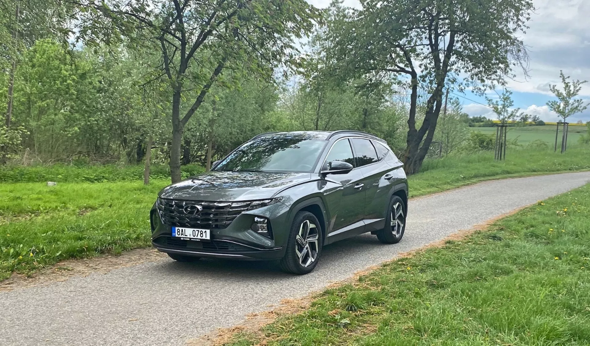 Vista frontal tres cuartos de un Hyundai Tucson 2020 crossover SUV estacionado en un camino pavimentado con vegetación de fondo, mostrando la parrilla distintiva del vehículo, faros LED elegantes, grandes ruedas de aleación de múltiples radios, línea de techo inclinada, alta distancia al suelo, espejos laterales integrados, vidrios polarizados y contornos estilizados del cuerpo.