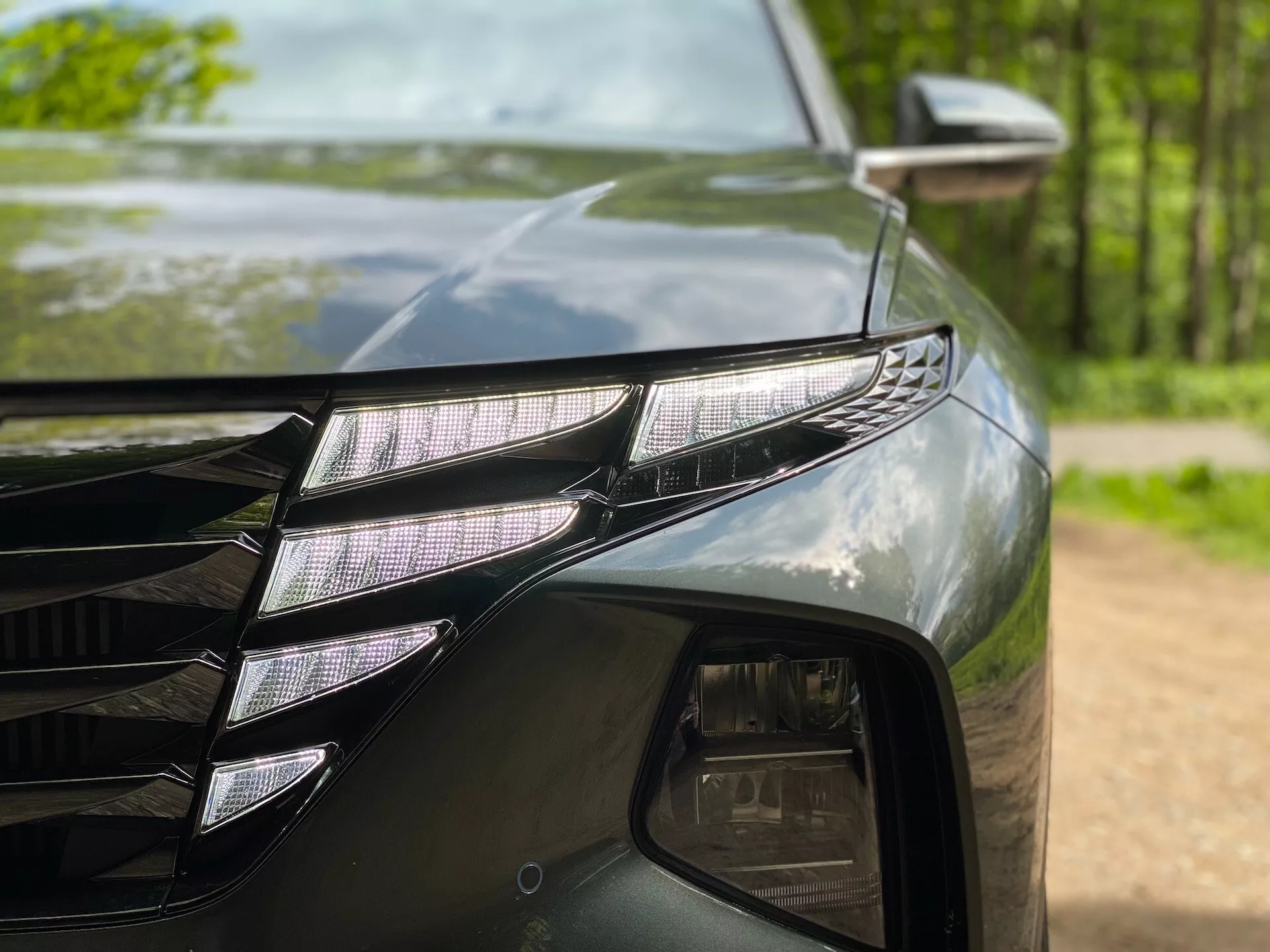 Primer plano del lado delantero izquierdo de un Hyundai Tucson 2020 (Hyundai-Tucson-1-6-T-GDi-Pure), mostrando un conjunto de faros LED con proyectores LED individuales y una tira DRL LED distintiva. La pintura oscura y brillante refleja los árboles circundantes. También se puede observar una parte de la parrilla con líneas angulares y el espejo lateral con un indicador de señal de giro.