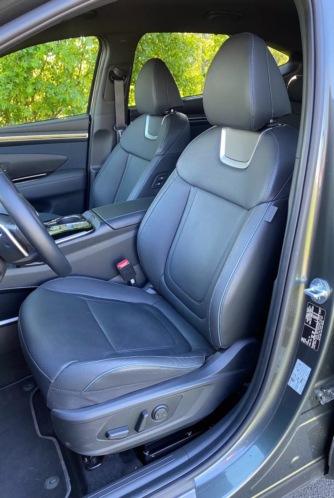 Interior del Hyundai Tucson 2020 1.6 T-GDI Pure que muestra el asiento delantero del pasajero con tapicería de cuero negro, costuras detalladas y controles de ajuste eléctrico. Consola central visible con portavasos, panel de puerta con materiales a juego, manija, compartimento de almacenamiento y rejilla del altavoz que sugiere un sistema de audio avanzado. Parte del tablero y la guantera parcialmente visibles. Alfombra tipo moqueta y leve reflejo de luz solar en el panel de la puerta.