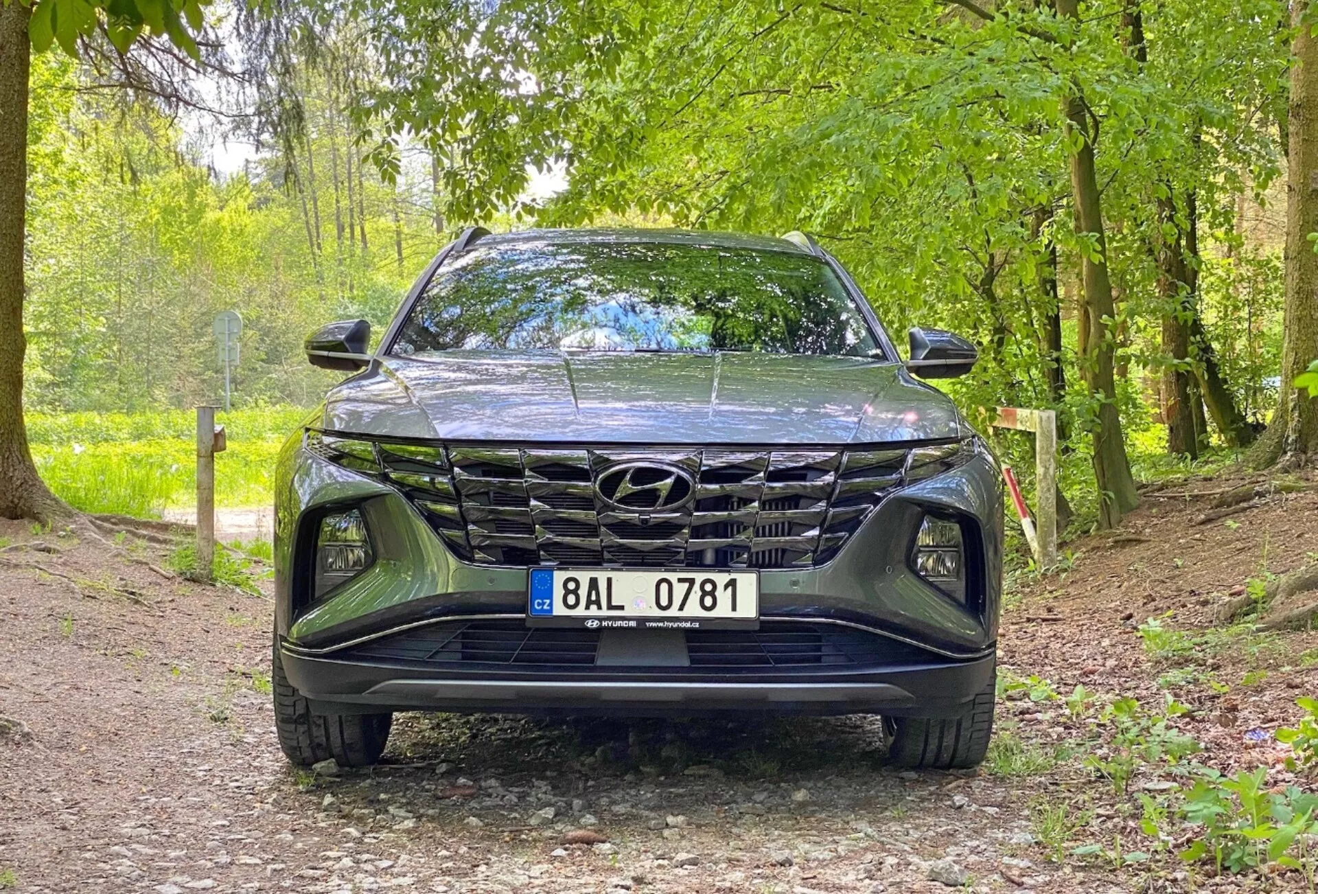 Vista frontal del vehículo Hyundai Tucson 1.6 T-GDi Pure del 2020, mostrando una toma en ángulo bajo centrada en la parrilla, el parachoques delantero y el área del capó. El coche está estacionado en un camino de grava en un entorno natural, lo que sugiere su idoneidad para entornos al aire libre. Las características clave incluyen un diseño de parrilla prominente, faros LED elegantes y el emblema de Hyundai visible.