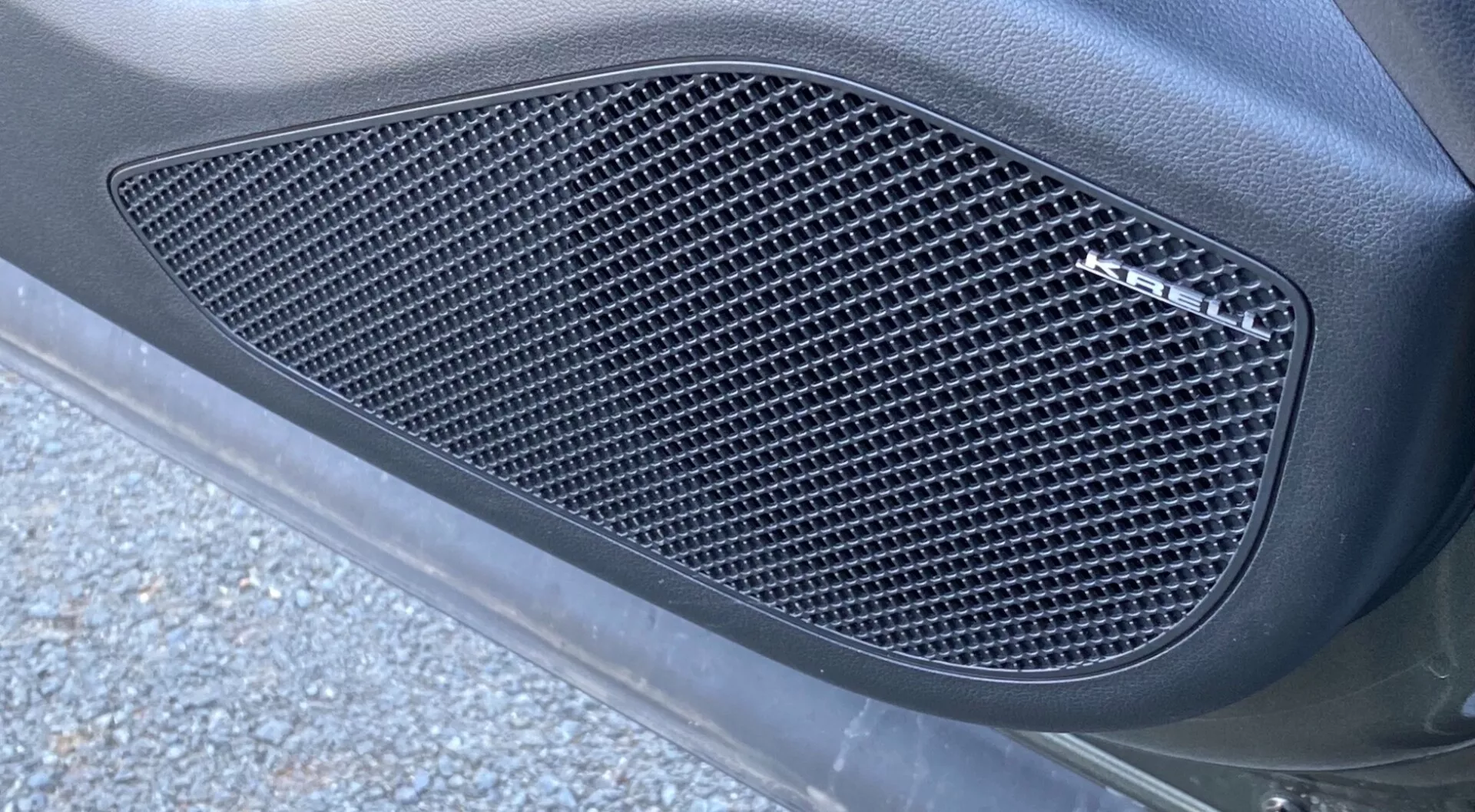 Vista lateral en primer plano del Hyundai Tucson 2020 1.6 T-GDI Pure, enfocándose en una rejilla lateral con un patrón de malla con recortes en forma de diamante. La rejilla, encajada en un contorno moldeado, está integrada en el panel de la carrocería del coche con un acabado mate, rodeada de una superficie curva y lisa. La imagen, capturada en ángulo oblicuo desde abajo, muestra iluminación natural al aire libre con pedazos de asfalto y escombros de la carretera, realzando la apariencia elegante y orientada al rendimiento.