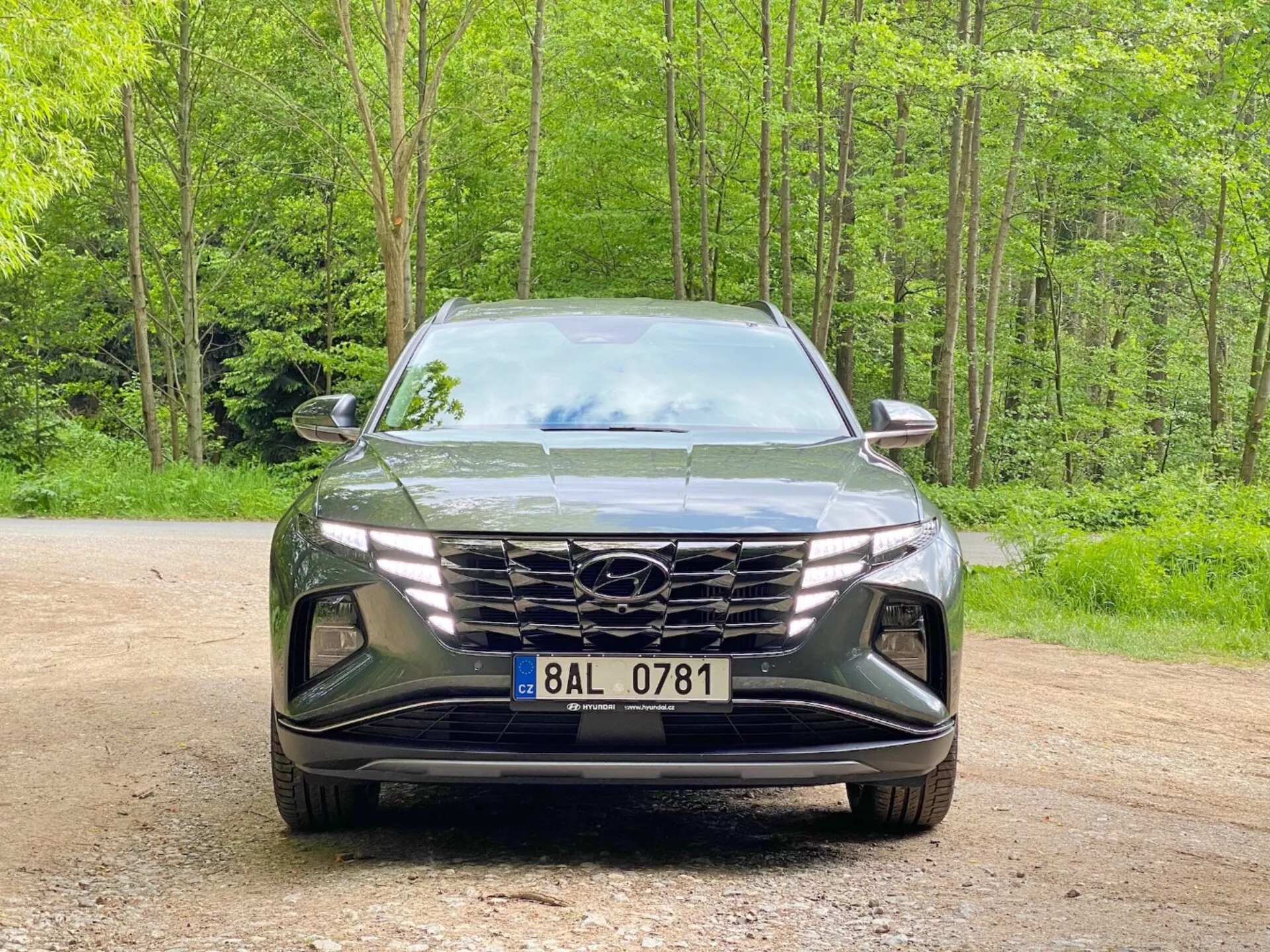 Vista frontal de un Hyundai Tucson 2020 (Hyundai Tucson 1.6 T-GDI Pure) SUV moderno estacionado en un camino de grava con un fondo boscoso. La imagen captura el estilo elegante y agresivo con una gran parrilla geométrica que presenta el logotipo del fabricante, faros LED angulares estrechos y un parachoques delantero esculpido con tomas de aire y huecos para luces antiniebla, destacando la avanzada aerodinámica y tecnología de iluminación.