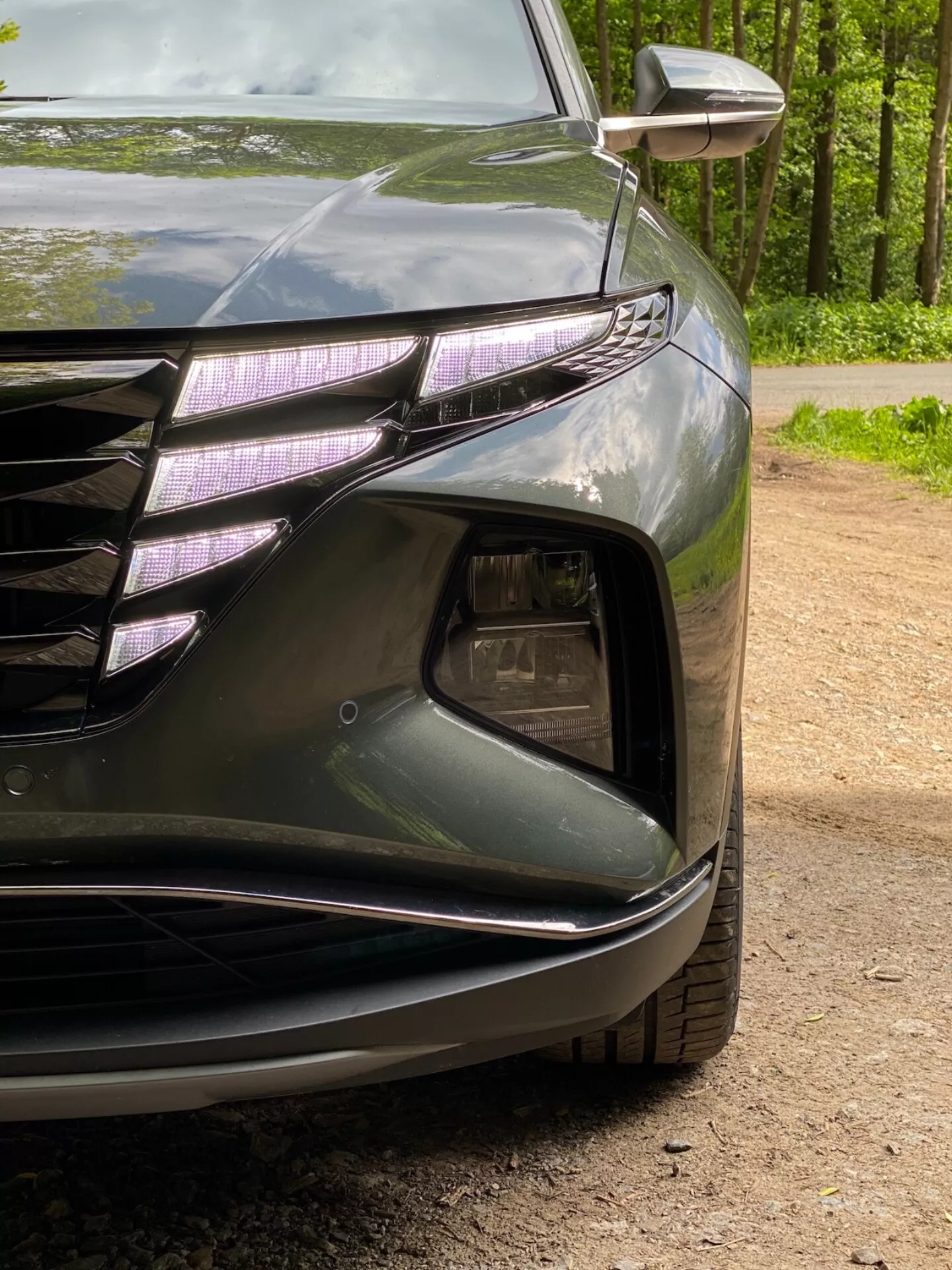 Vista de cerca de la esquina delantera izquierda de un Hyundai Tucson 2020 1.6 T-GDI Pure, que muestra el faro con luces diurnas LED y lente tipo proyector, parte de la parrilla delantera con líneas horizontales y acentos cromados, sección de entrada de aire con alojamiento para faros antiniebla, parachoques delantero esculpido con líneas suaves y señal de giro integrada en el espejo exterior. La pintura oscura y brillante resalta las curvas aerodinámicas del coche.