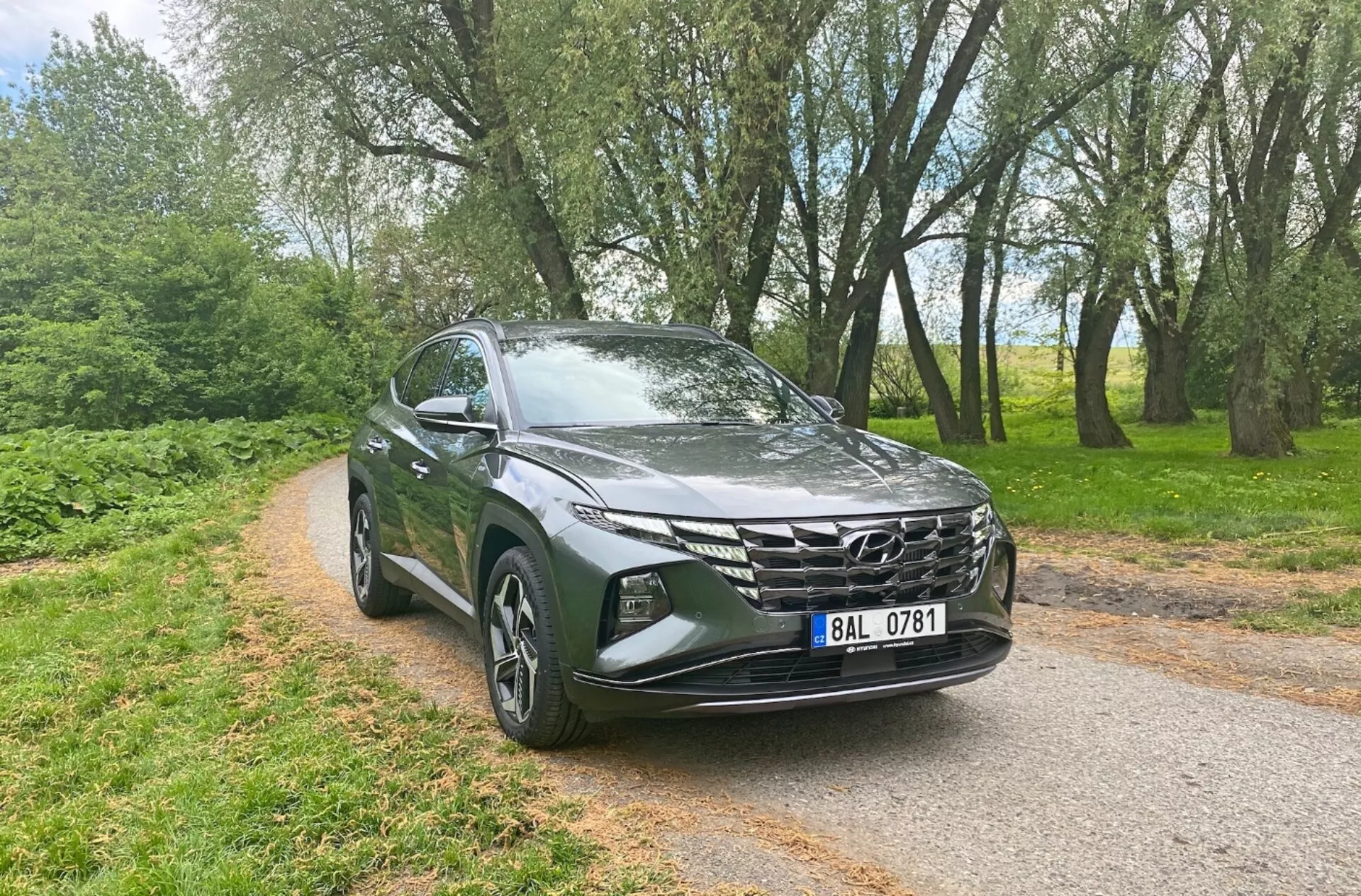 Vista tres cuartos frontal de un Hyundai Tucson 1.6 T-GDi Pure 2020, que muestra características modernas de SUV como faros LED elegantes, una parrilla delantera geométrica prominente, capó aerodinámico esculpido, llantas de aleación, espejos laterales con indicadores de señal, y pintura metálica, ambientado en un camino pavimentado estrecho con árboles de fondo, resaltando su versatilidad y atractivo para entornos urbanos y rurales.