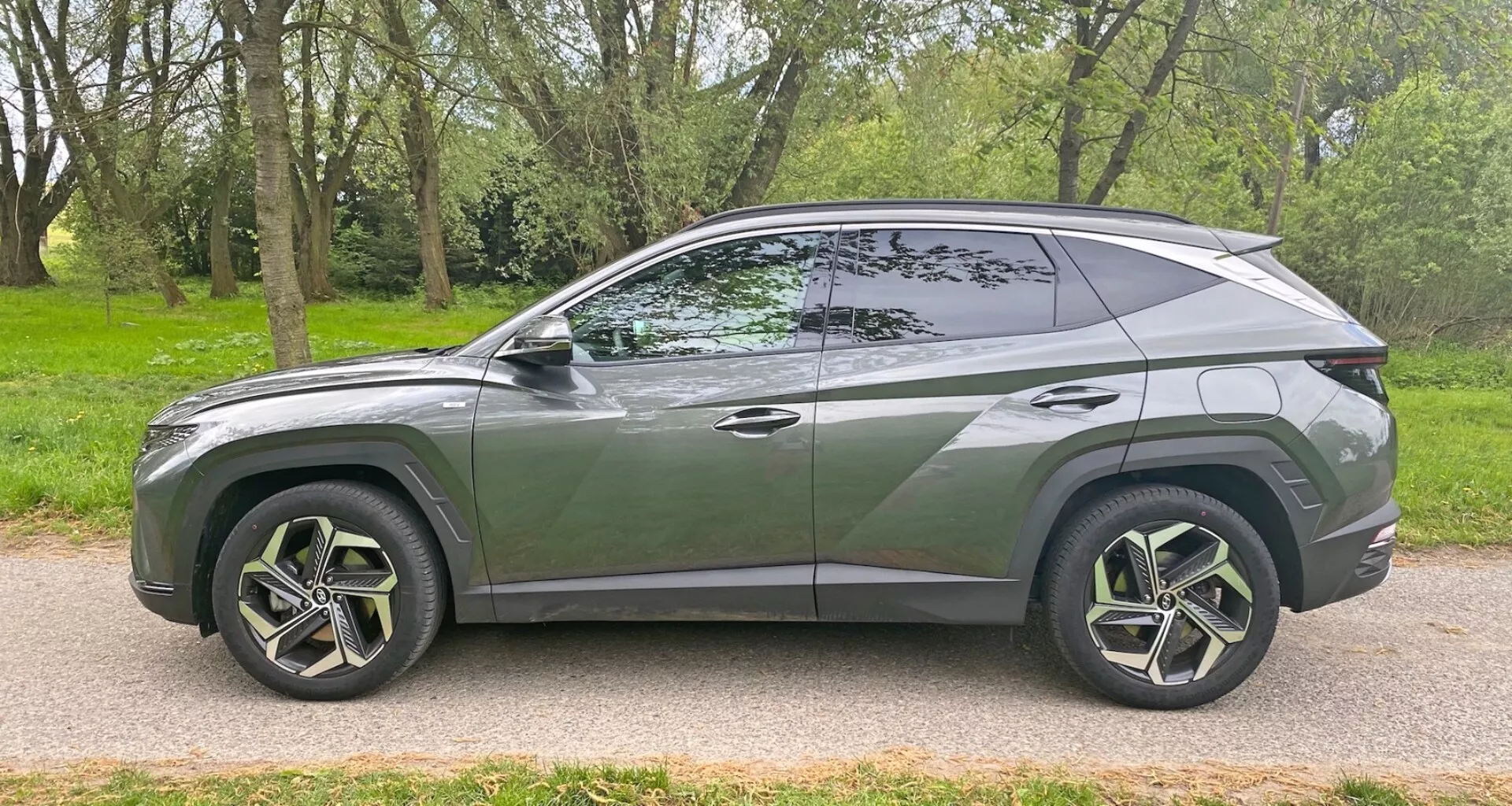 Perfil lateral del moderno SUV Hyundai Tucson 1.6 T-GDI Pure 2020 estacionado en una carretera pavimentada junto a una zona de césped con árboles. Presenta un diseño aerodinámico y elegante con una línea de techo descendente, una línea de carrocería distintiva, grandes llantas de aleación con múltiples radios y neumáticos de perfil bajo, ventanas tintadas con un pilar B visible, manijas de puertas al ras, espejos laterales del color de la carrocería y molduras cromadas a lo largo del borde de la ventana. El vehículo captura un estilo contemporáneo y posibles características avanzadas desde un ángulo lateral.