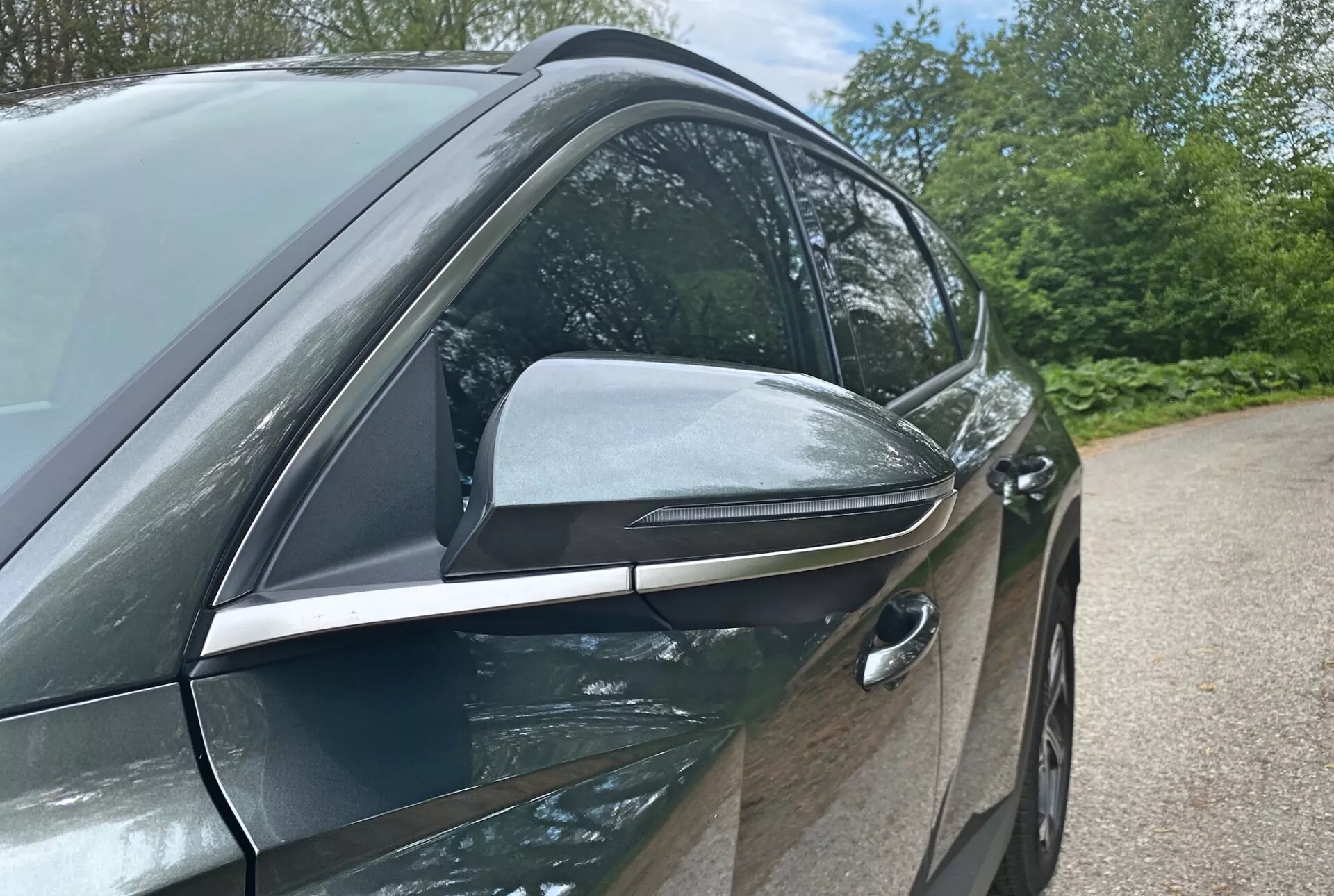 Vista lateral de cerca del Hyundai Tucson 2020 1.6 T-GDI Pure que destaca su diseño moderno y características tecnológicas con enfoque en el lado del conductor. La fotografía en ángulo bajo resalta el espejo lateral aerodinámico con indicadores de señal de giro integrados y acentos negros, sugiriendo funcionalidades avanzadas. La manija de puerta delantera del conductor, empotrada, indica un sistema de entrada sin llave, contribuyendo a la eficiencia aerodinámica del vehículo. Las puertas de color de carrocería brillante y las molduras de cromo en las ventanas sugieren un paquete de acabados premium. Elementos de fondo como árboles y cielo sugieren un entorno al aire libre.