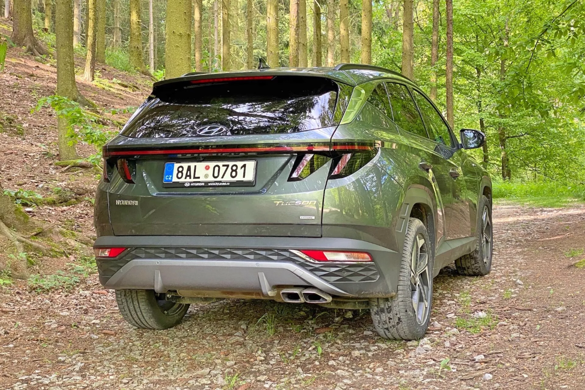 Imagen de un moderno SUV Hyundai Tucson 2020 en un entorno forestal con camino de grava, capturada desde un ángulo trasero de tres cuartos. La foto destaca el lado derecho y el perfil trasero del vehículo con el portón trasero visible, luces traseras LED, sistema de escape doble, alerón trasero y llantas de aleación.