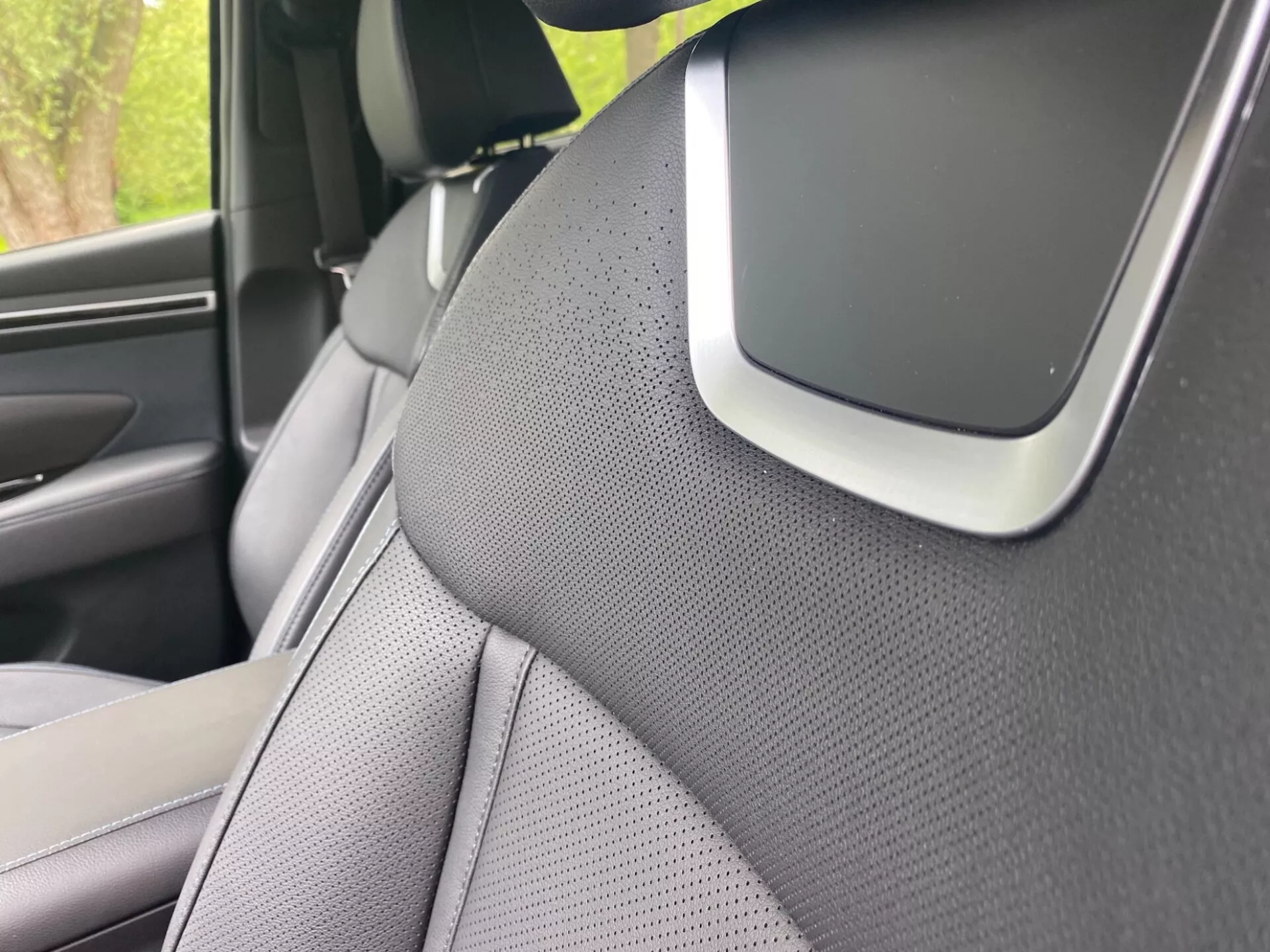 Primer plano del interior del Hyundai Tucson 2020 centrado en el pilar A derecho y parte del asiento delantero derecho. El pilar A está cubierto con un material texturizado, probablemente tela o cuero sintético, sugiriendo un enfoque estético o de reducción de ruido. El asiento tiene un diseño bicolor y está perforado para ventilación. Una característica notable en la unión del pilar A y el tapizado del techo podría ser parte del sistema de audio, un componente de airbag o un sensor de asistencia al conductor, destacando características avanzadas de seguridad o tecnología.