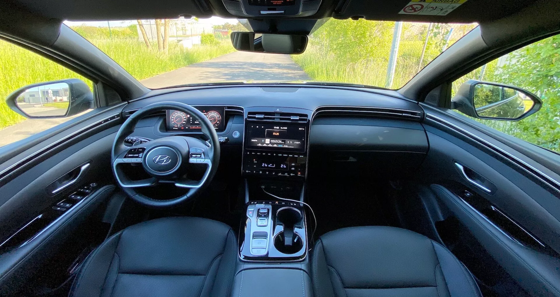 Vista del interior del Hyundai Tucson 2020 desde el asiento trasero, mostrando el panel de control frontal, volante con controles integrados, cuadro de instrumentos digital, gran sistema de infoentretenimiento con pantalla táctil, botones de control de clima, palanca de cambios y asientos delanteros tapizados en cuero bajo la iluminación natural del día.