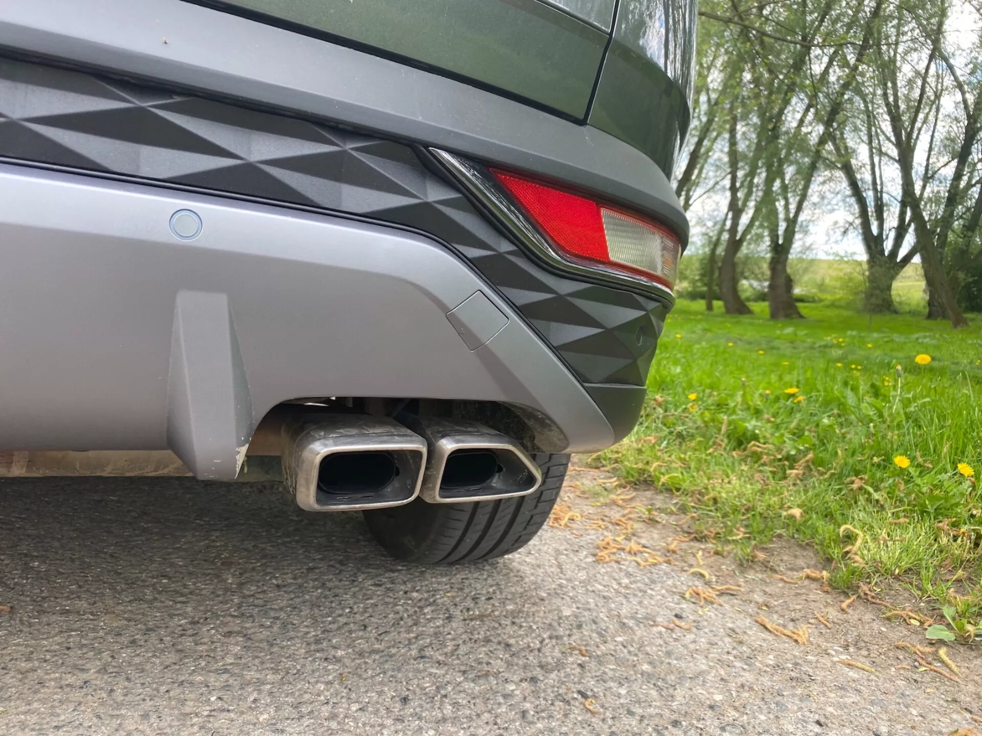 Imagen de primer plano de la sección trasera de un Hyundai Tucson 2020, con un ángulo a nivel del suelo. Los detalles visibles incluyen un parachoques trasero de dos tonos, una luz trasera LED parcial y tubos de escape dobles de metal con puntas rectangulares, que sugieren un acabado deportivo. El diseño elegante del vehículo y la pintura metálica reflejan el entorno natural.