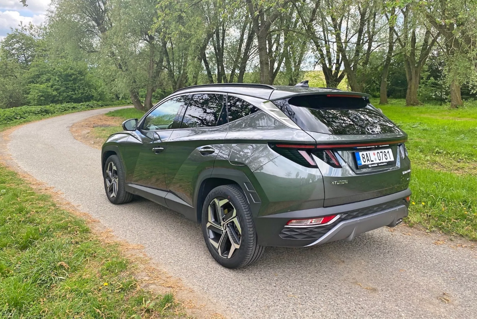 La imagen muestra un SUV crossover moderno desde un ángulo trasero de tres cuartos, destacando el diseño aerodinámico y la línea de techo inclinada. El vehículo verde oscuro presenta una puerta trasera estilo liftback, un alerón deportivo y grandes ruedas de aleación con diseño de múltiples radios. Los elementos visibles incluyen luces traseras LED modernas, salidas de escape dobles y un limpiaparabrisas trasero. El SUV está estacionado en un camino de grava rodeado de vegetación, lo que sugiere un entorno de parque o rural.
