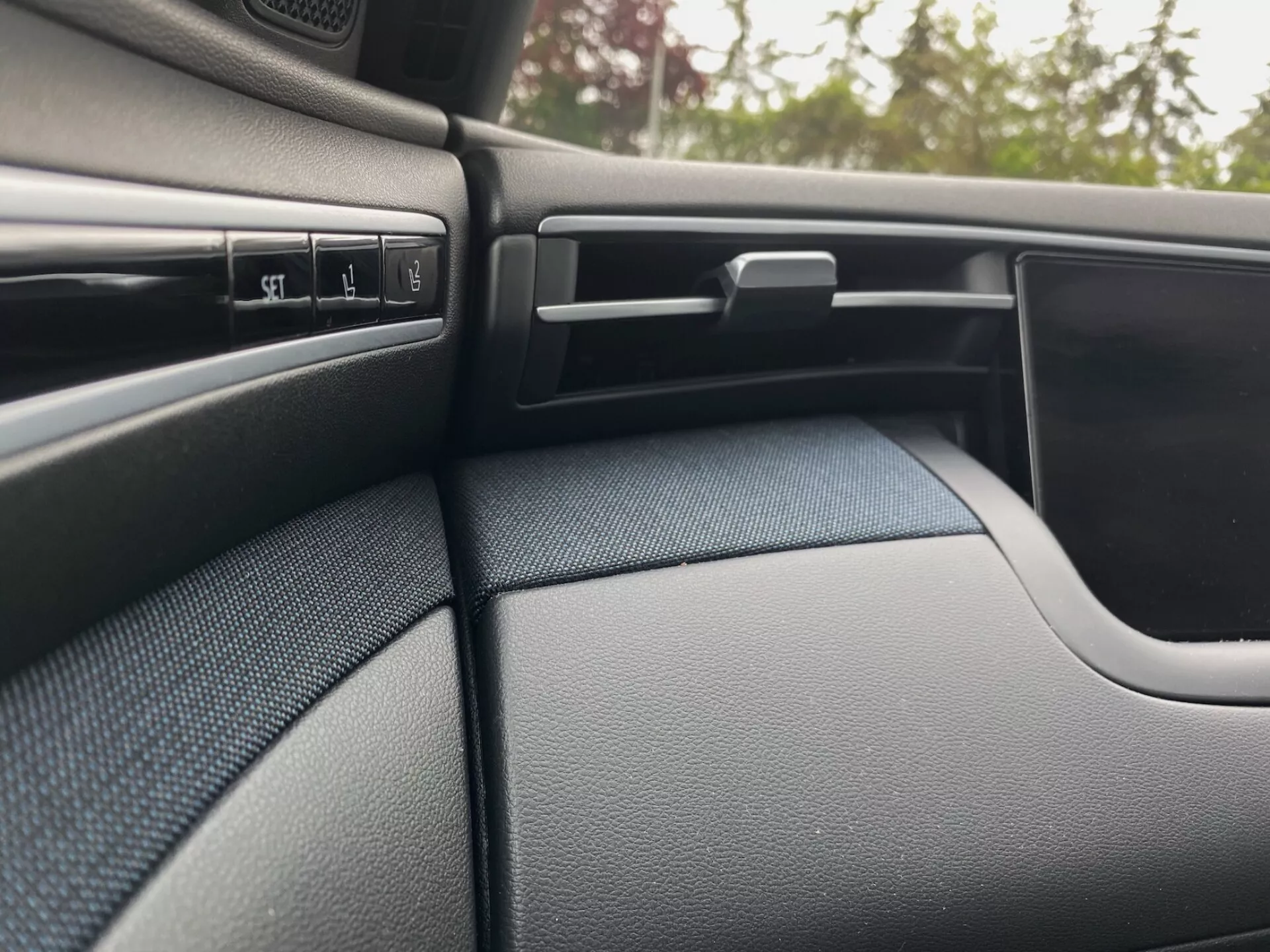 Detalle interior del Hyundai Tucson 2020, mostrando el salpicadero texturizado de color oscuro del lado del pasajero y parte del panel de la puerta. La escena resalta varios botones de control, una salida de aire ajustable con lamas horizontales, una guantera con cerradura y el diseño curvo del panel de la puerta que se integra perfectamente con el salpicadero.