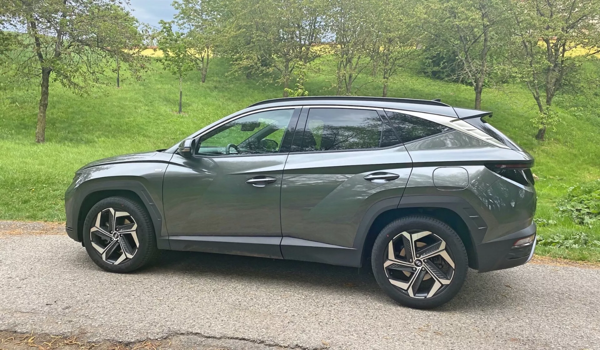 Vista de perfil lateral del Hyundai Tucson 1.6 T-GDI Pure 2020 SUV en un entorno natural con luz diurna. El vehículo está ubicado en una zona de césped con árboles al fondo, sugiriendo un entorno rural o suburbano. Características de diseño moderno incluyen faros LED, señales de giro integradas en los espejos laterales, ventanas tintadas, ruedas de aleación aerodinámicas y una línea de techo inclinada estilo cupé. La carrocería elegante con líneas de carácter y manijas de puertas empotradas sugieren un sistema de entrada sin llave avanzado, mientras que el alerón trasero indica mejoras aerodinámicas. El coche está ubicado centrado en el marco, proporcionando una vista clara y despejada desde una perspectiva de altura media.