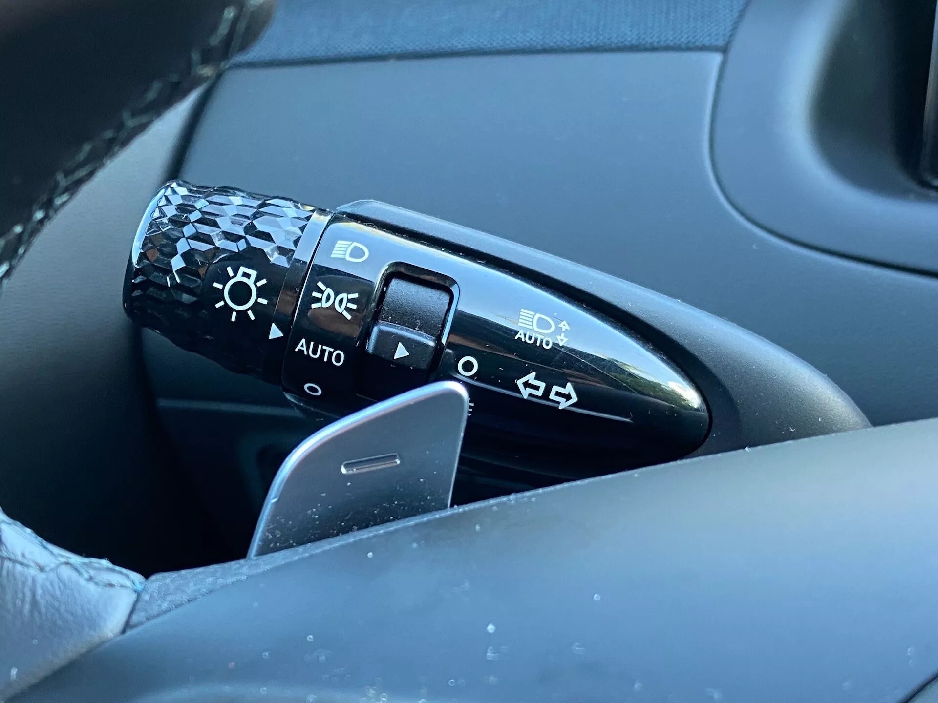 Vista de primer plano de la palanca de control ubicada en la columna de dirección a la izquierda del volante en un Hyundai Tucson 2020. La palanca presenta símbolos para configuraciones de faros incluidos apagado, faros automáticos, luces de estacionamiento y faros de luz baja/alta. La imagen captura la palanca, parte del volante y una porción de la puerta del lado del conductor en un fondo desenfocado, enfocándose en el detalle y la calidad del diseño. La posición 'AUTO' indica un sistema de faros automáticos.