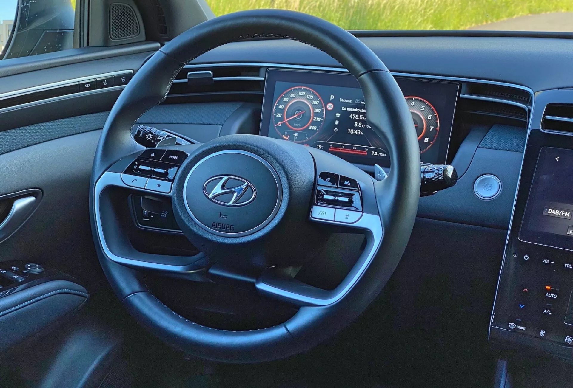 Vista interior del Hyundai Tucson 1.6 T-GDI 2020 desde la perspectiva del conductor, enfocando el volante de tres radios moderno con el logotipo de Hyundai, botones de control de medios y crucero, tablero de instrumentos parcialmente visible con velocímetro y cuentarrevoluciones, y sistema de infoentretenimiento con pantalla táctil central en el tablero.