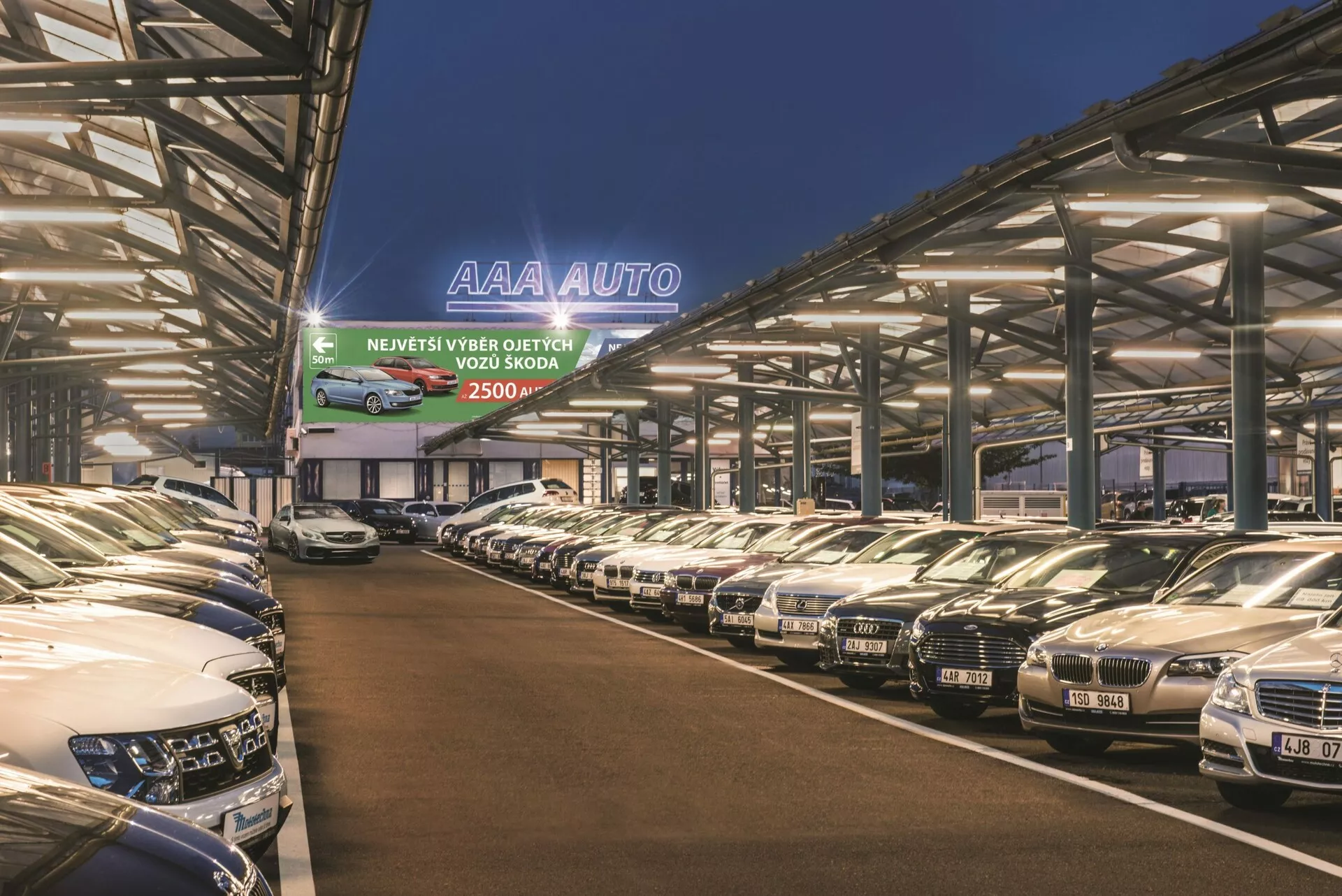 Hyundai Tucson 2018 expuesto en un lote de concesionario AAA AUTO durante el crepúsculo con diversos modelos iluminados por luces artificiales, revelando sus parrillas y faros bajo una estructura metálica protectora