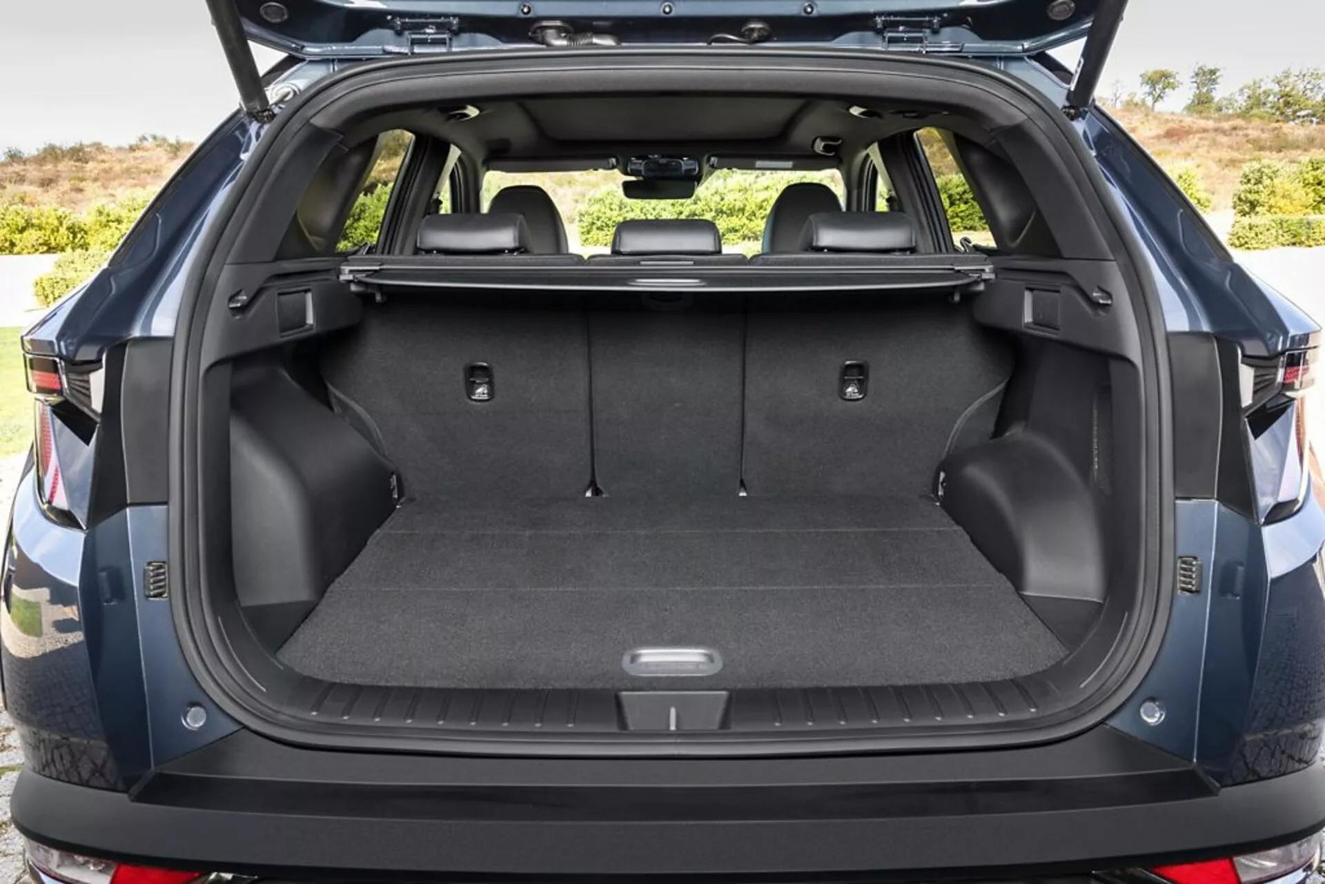 Área de carga trasera de Hyundai Tucson 2020 con maletero abierto, mostrando suelo de carga alfombrado, puntos de anclaje, compartimientos de almacenaje, toma de 12V, asientos traseros plegables, cubierta de carga retráctil, paredes laterales de plástico duradero, y un vistazo al parachoques trasero, en un entorno al aire libre.