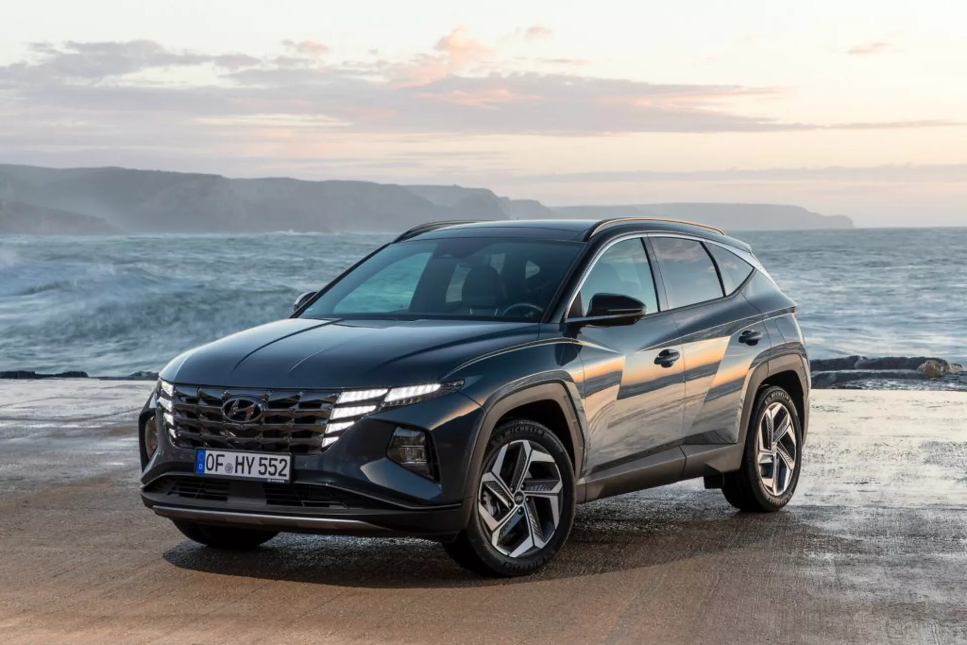 SUV moderno Hyundai Tucson 2020 estacionado frente a la playa con el horizonte del océano de fondo, mostrando su vista frontal de tres cuartos que destaca el diseño del vehículo, la parrilla prominente con logo, faros LED angulares y ruedas de aleación deportivas