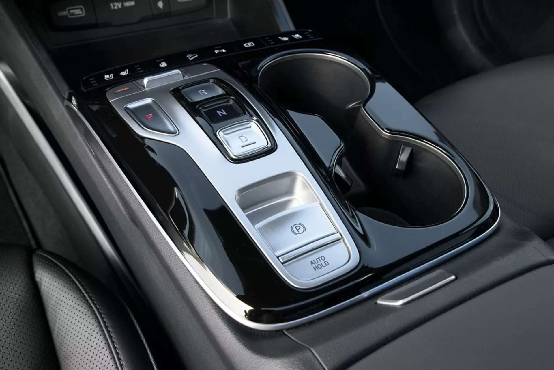 Vista cercana de la consola central del Hyundai Tucson 2020 mostrando la palanca de cambios automática, botones de control, función Auto Start/Stop, interruptor de luces de emergencia y porta-vasos doble con detalles cromados, desde una vista superior con ligera inclinación.