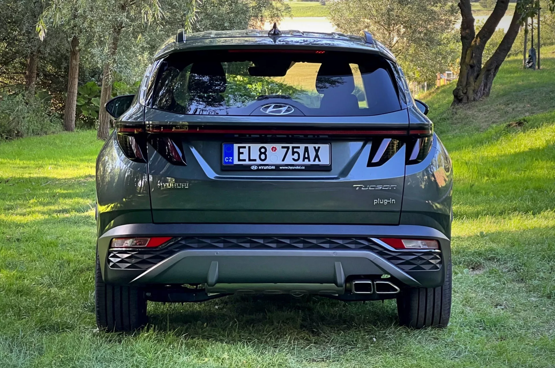 Vista trasera del SUV Hyundai Tucson 2020 en pendiente herbosa con fondo de bosque, mostrando logo en portón trasero, insignias Tucson y Hybrid, señal de Plug-in, luces reflectoras, luces traseras dinámicas, limpiaparabrisas trasero, luz de freno montada alto, puntas de escape dobles, placas de deslizamiento plateadas y distintivo AWD.