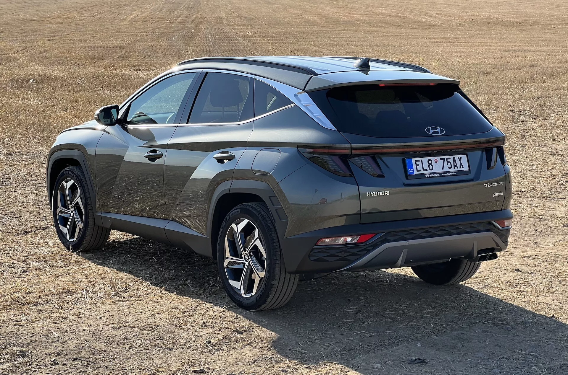 SUV moderno Hyundai Tucson 2020 en vista oblicua mostrando perfiles traseros y laterales con distintivo de tecnología híbrida en campo herboso seco, destacando luces traseras LED, inserción de parachoques contrastante y ruedas deportivas de múltiples radios
