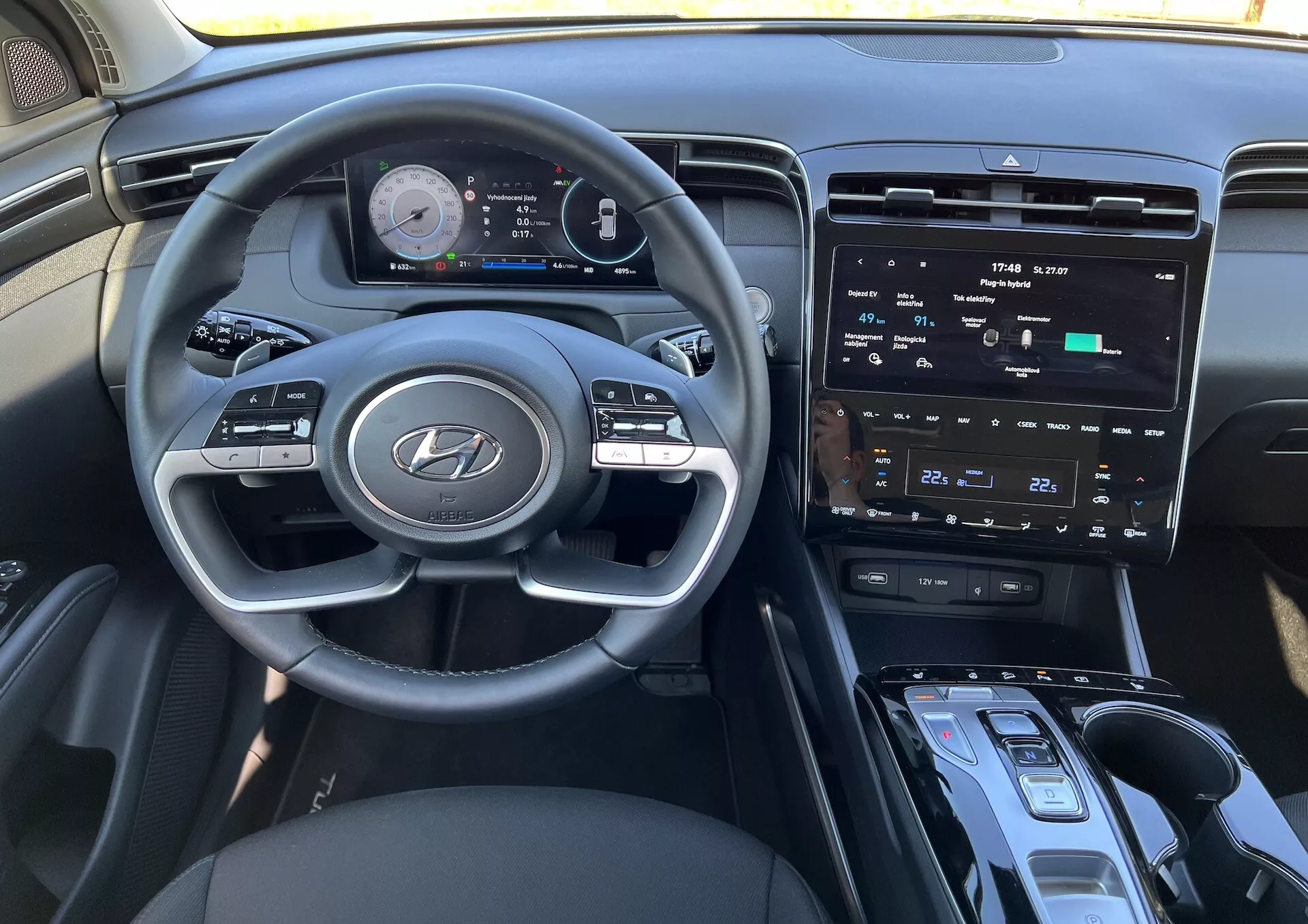 Interior del Hyundai Tucson 2020 mostrando el volante con logo de Hyundai, cuadro de instrumentos digital avanzado, pantalla táctil central de infoentretenimiento, controles de clima, botón de encendido/apagado, palanca de cambios, y posible área de carga inalámbrica desde la perspectiva del conductor