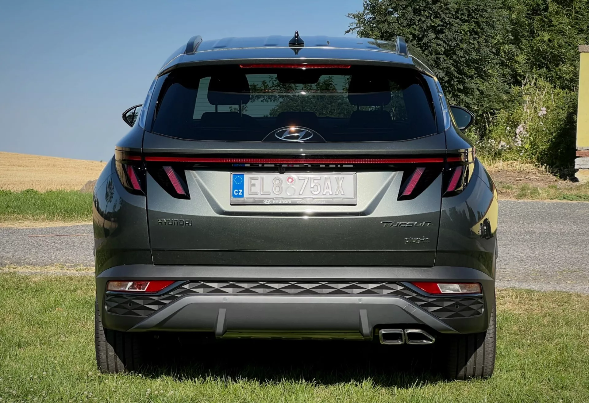 Vista trasera del Hyundai Tucson 2020 mostrando luces traseras horizontales, insignia central, nombre del nivel de acabado, sensores de aparcamiento integrados en el parachoques y sistema de escape doble, aparcado en una superficie bordeada de césped contra un cielo despejado