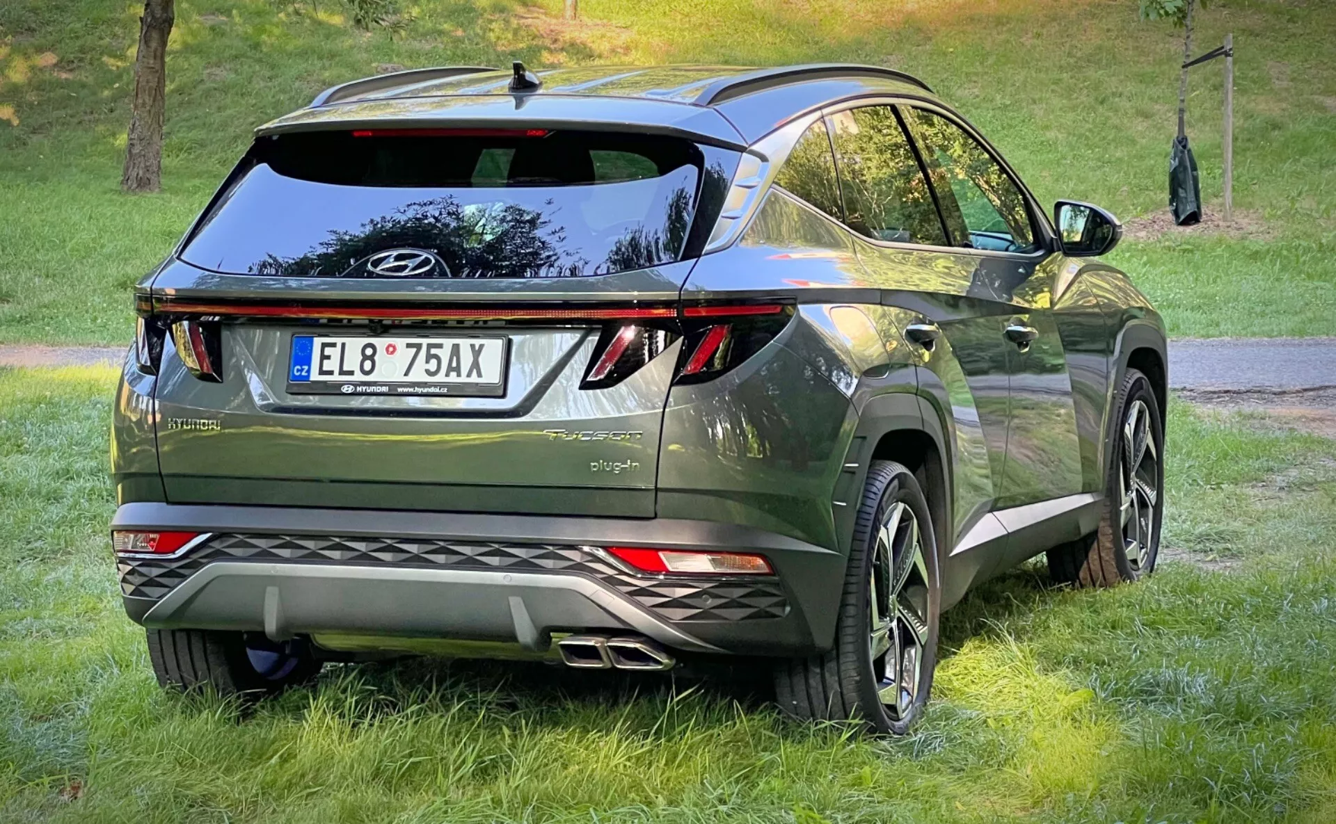 Vista trasera tres cuartos de un Hyundai Tucson 2020, mostrando sus luces traseras LED, diseño aerodinámico con spoiler en el techo y antena de aleta de tiburón, acabado premium en verde metálico oscuro, y equipado con grandes llantas de aleación y sensores de estacionamiento, en un entorno de estilo de vida crossover urbano