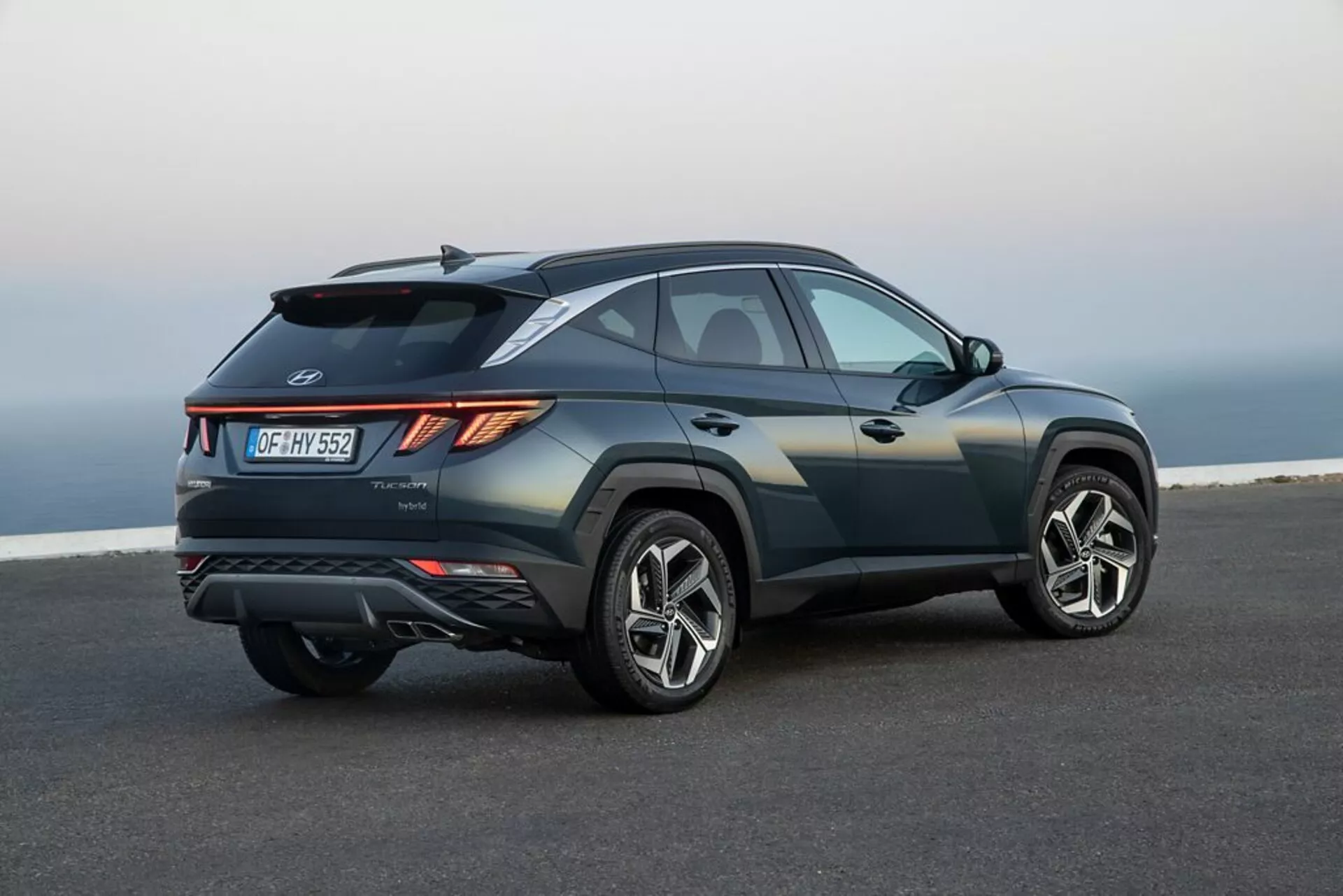 SUV Hyundai Tucson 2020 estacionado en asfalto liso con un paisaje costero o elevado al fondo, mostrando su diseño elegante y deportivo, línea de cintura ascendente, línea de techo inclinada, pliegues afilados del cuerpo, luces traseras LED angulares, spoiler trasero, difusor integrado, doble escape y grandes ruedas de aleación