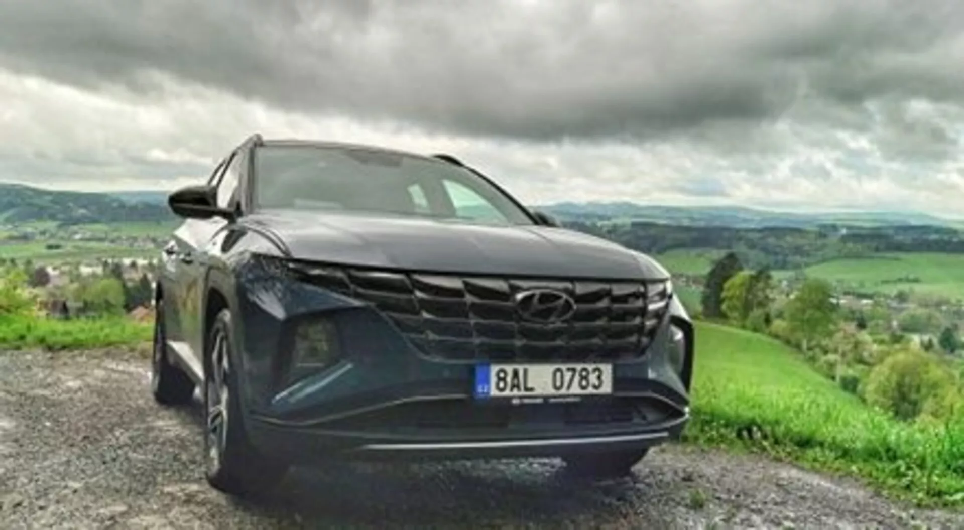 SUV Hyundai Tucson 2020 estacionado en un camino de grava, mostrando su audaz parrilla, elegantes faros LED y pintura metálica en una vista dinámica desde un ángulo frontal bajo, con un paisaje montañoso y exuberante bajo un cielo nublado al fondo