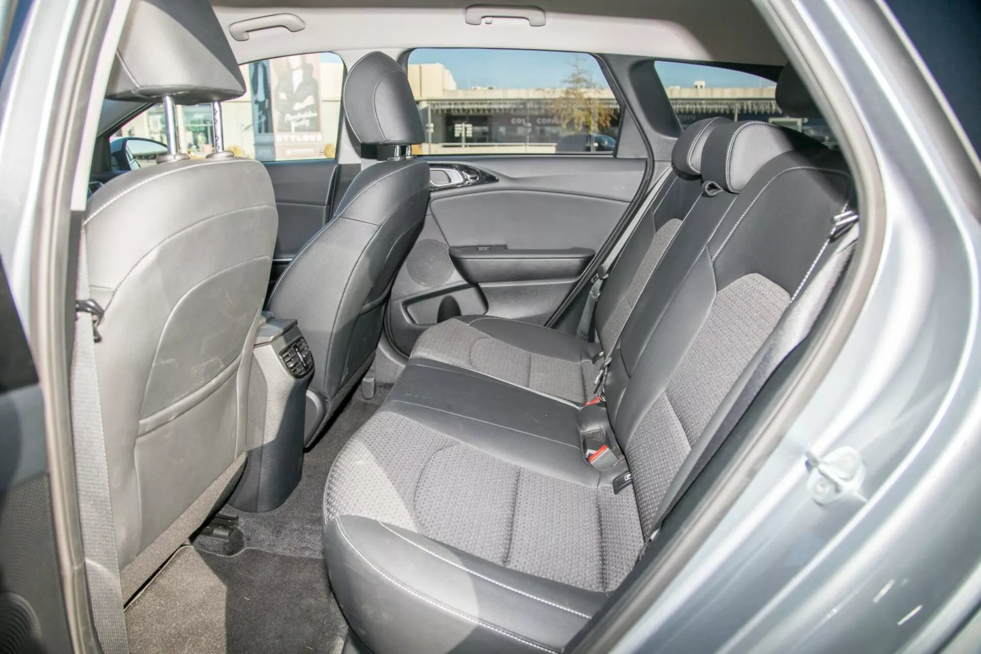 Vista interior de un Kia Cee'd 2009 mostrando el diseño y las características del área de asientos traseros, destacando la tapicería de tela duradera en dos tonos, cinturones de seguridad visibles, reposacabezas ajustables, panel de puerta con compartimiento de almacenamiento, posibles anclajes para asientos de niños, suelo enmoquetado y iluminación ambiental domo desde la perspectiva de la puerta trasera lateral derecha.