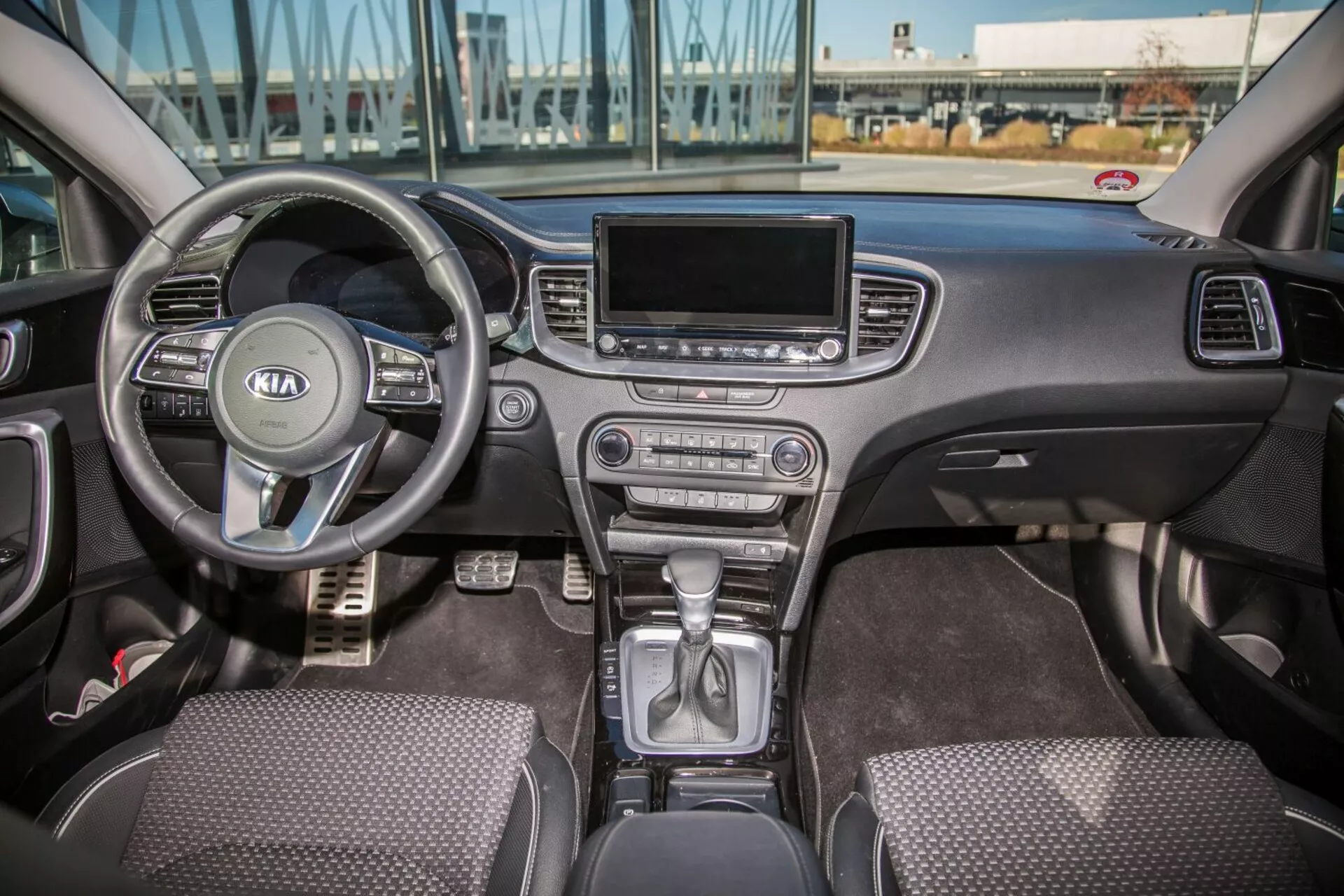 Vista interior del Kia Cee'd 2009 mostrando la cabina frontal completa incluyendo el volante multifunción con logo de Kia, cluster de instrumentos detrás, palanca de cambios en posición de park en la consola central, botones de control climático, pantalla de infotenimiento, asientos con tapizado de tela y controles de puertas de conductor y pasajero en luz natural diurna