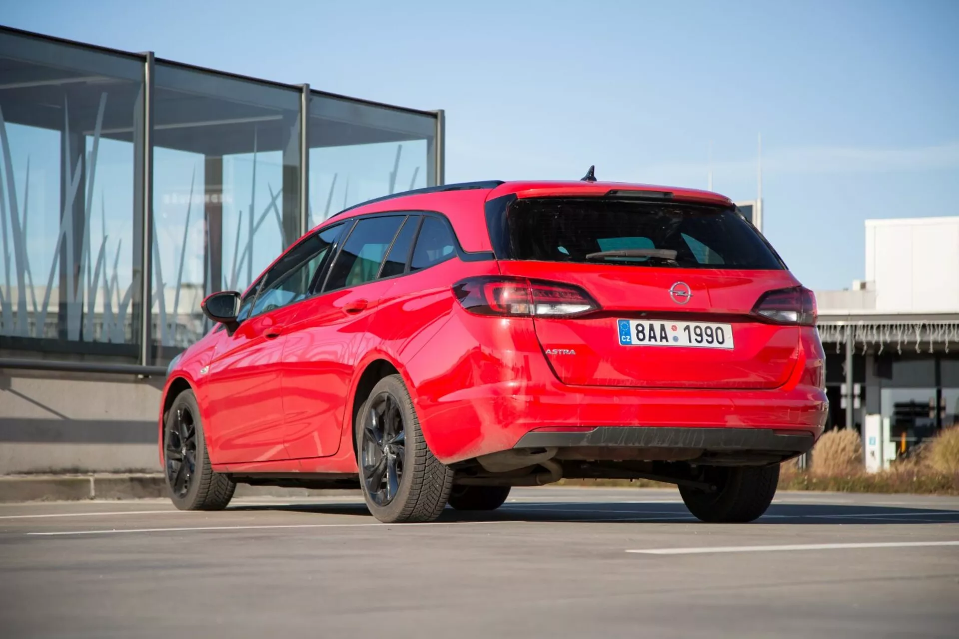 Kia Ceed hatchback 2009 en rojo mostrando su vista trasera deportiva de tres cuartos sobre concreto con edificio moderno de fachada de vidrio al fondo, exhibiendo diseño de luces traseras distintivo, spoiler de techo, antena aleta de tiburón, llantas de aleación y posibles sensores de estacionamiento.