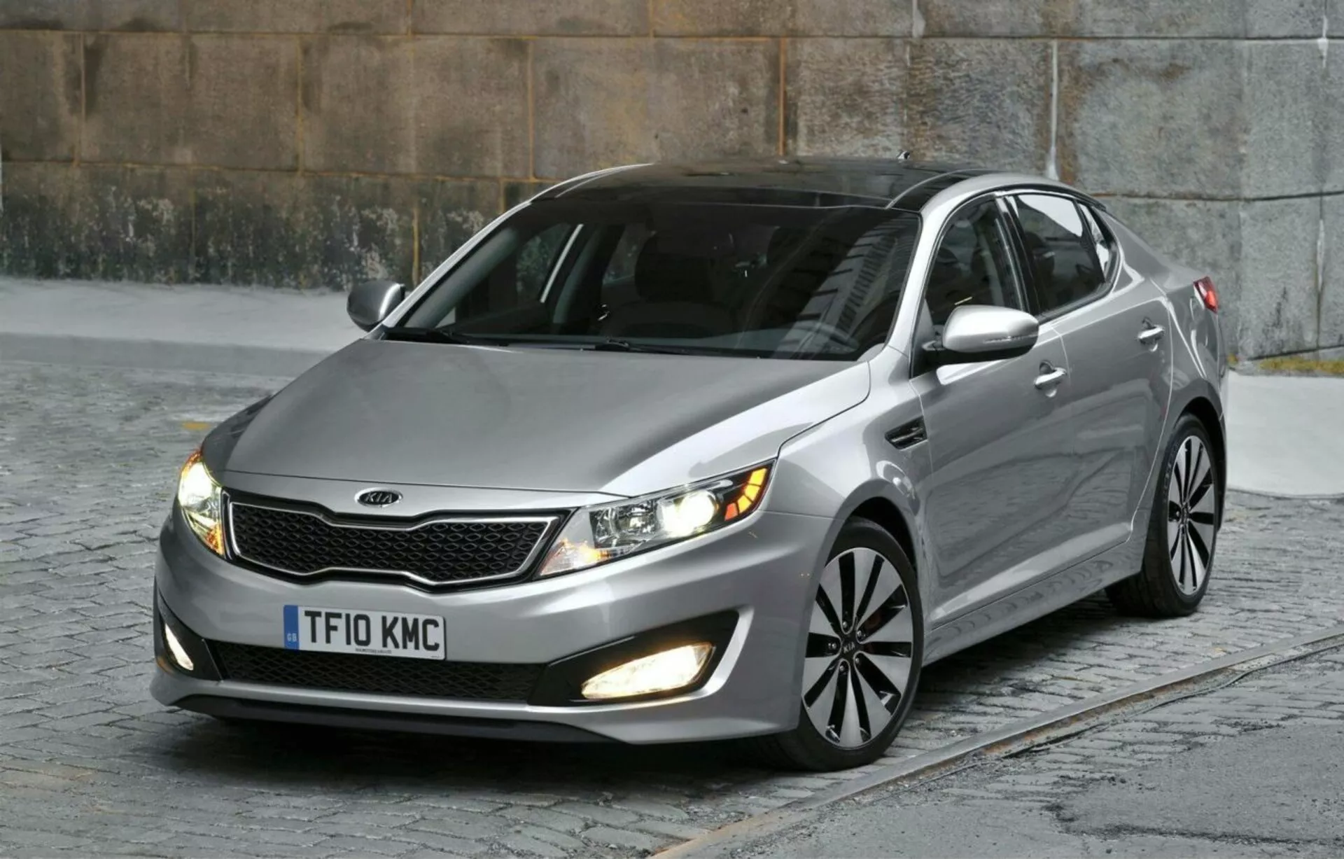 Kia Optima Limusina 2012 aparcado sobre adoquines capturando su cuarto frontal izquierdo incluyendo la parrilla con acentos cromados, faros con lentes claros, luces antiniebla, pintura metálica, llantas de aleación multirradio e indicadores de señal de giro integrados, enfatizando su diseño aerodinámico y características de sedán moderno en un entorno urbano