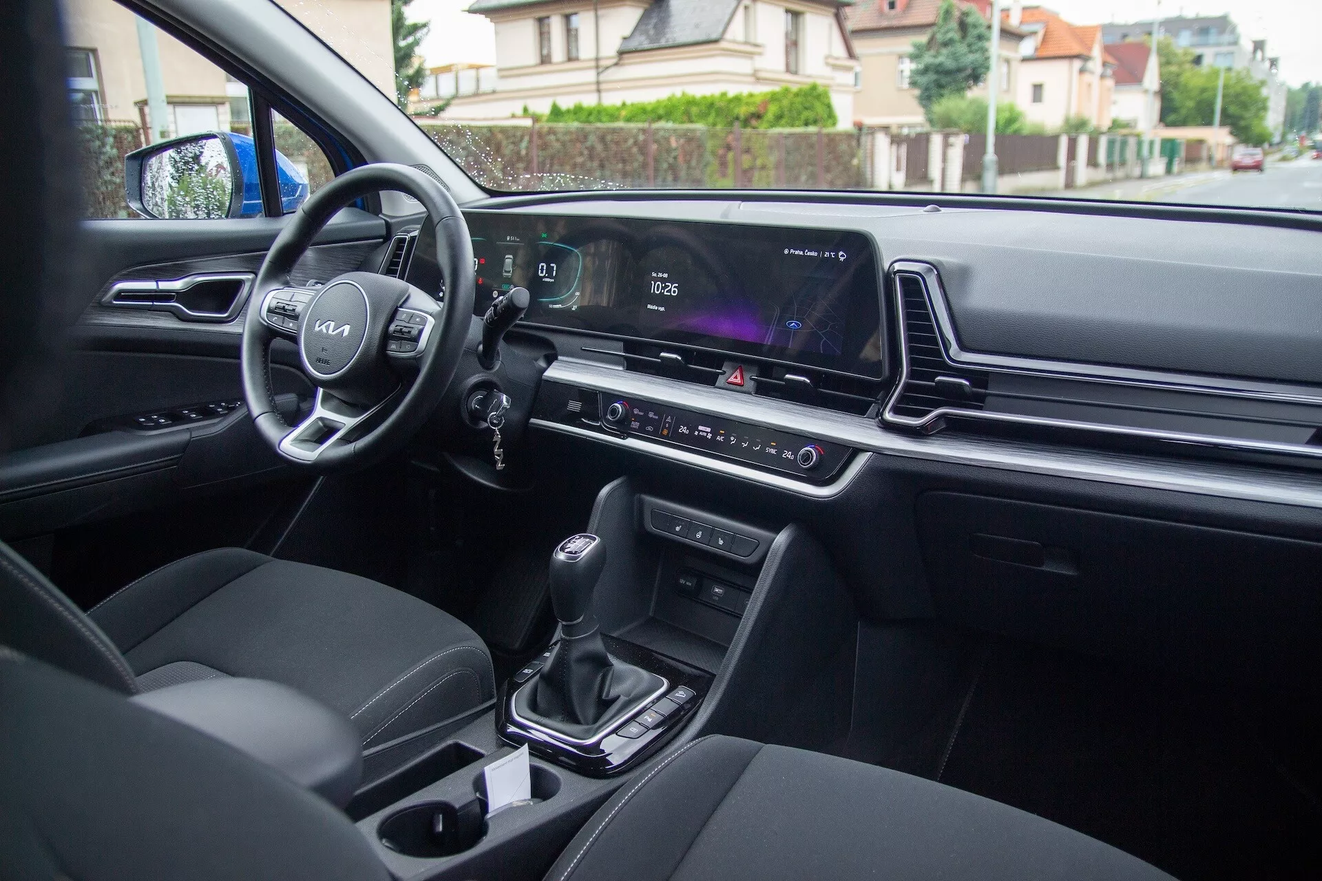 Vista interior del Kia Sportage 2022 desde el asiento trasero mostrando controles en la puerta del conductor, volante con logo y controles montados, panel de instrumentos digital, pantalla de infotenimiento, control de clima, palanca de cambios automática, portavasos y asientos tapizados modernos