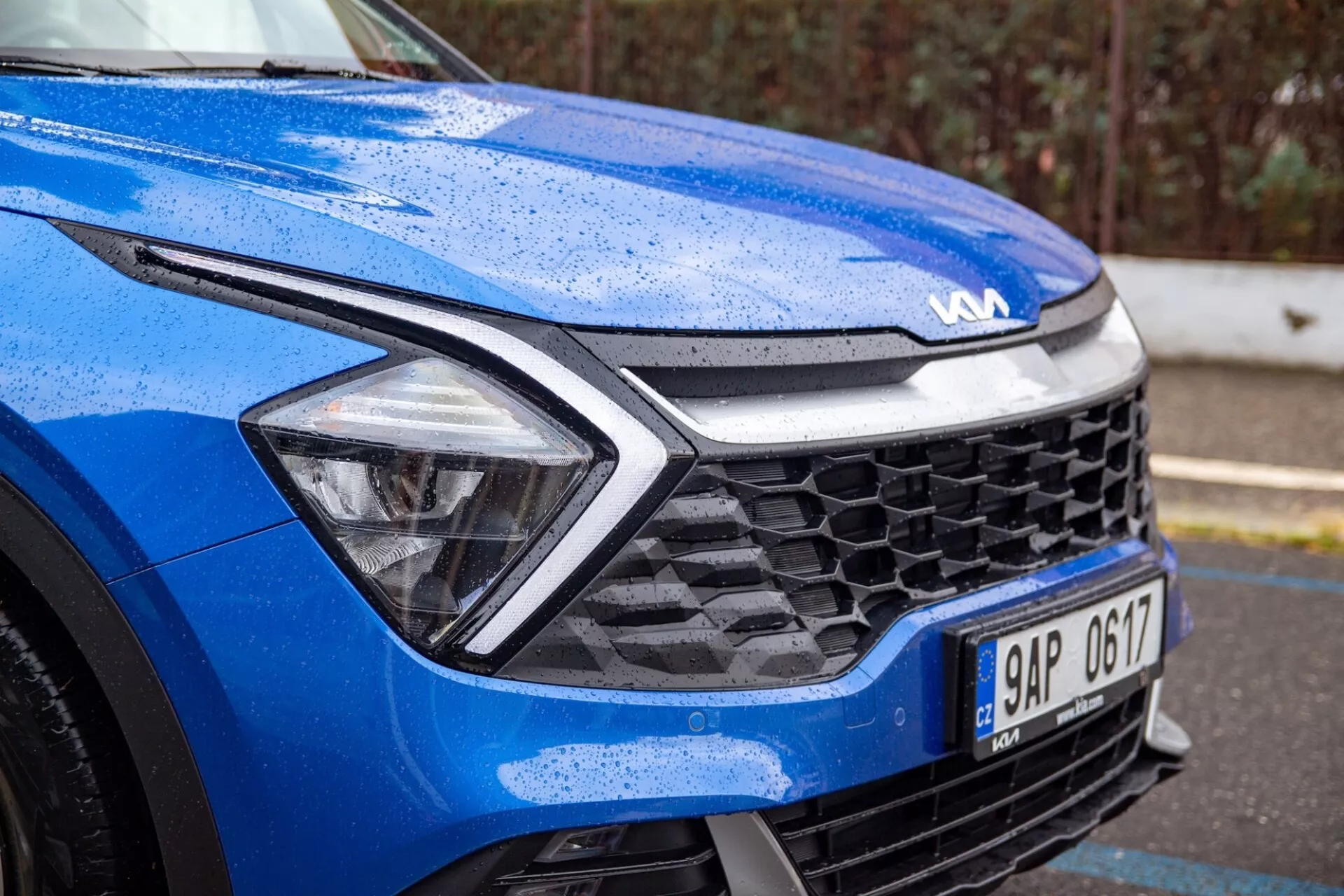 Vista detallada de un Kia Sportage azul 2022 centrado en el cuarto delantero izquierdo bajo condiciones húmedas, destacando el diseño dinámico del capó, faros LED, parrilla de panal y tecnología de sensores avanzada