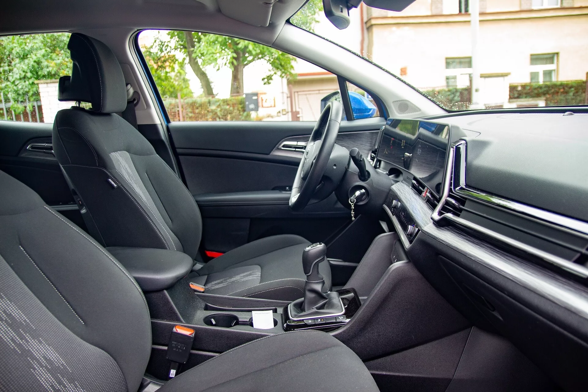 Vista interior del Kia Sportage 2022 desde la perspectiva del pasajero trasero mostrando el asiento del pasajero delantero, la consola central con cambio de marchas automático, el volante a la izquierda y una gran pantalla de infoentretenimiento en el tablero