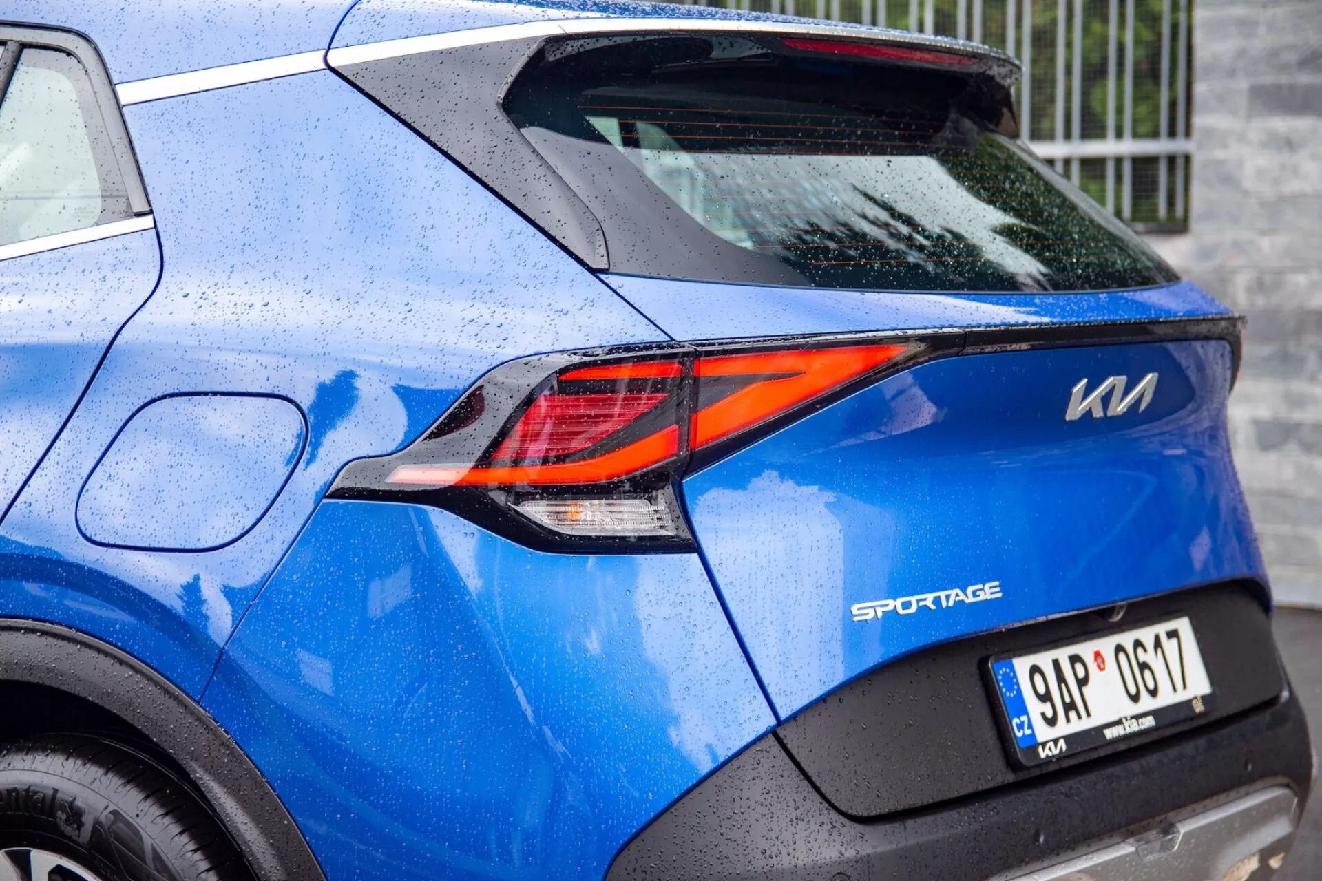 Vista parcial de un Kia Sportage azul 2022 que destaca la sofisticada luz trasera LED en forma de Z y los detalles del diseño trasero, incluido el distintivo del modelo, la placa de matrícula y la pintura salpicada de lluvia