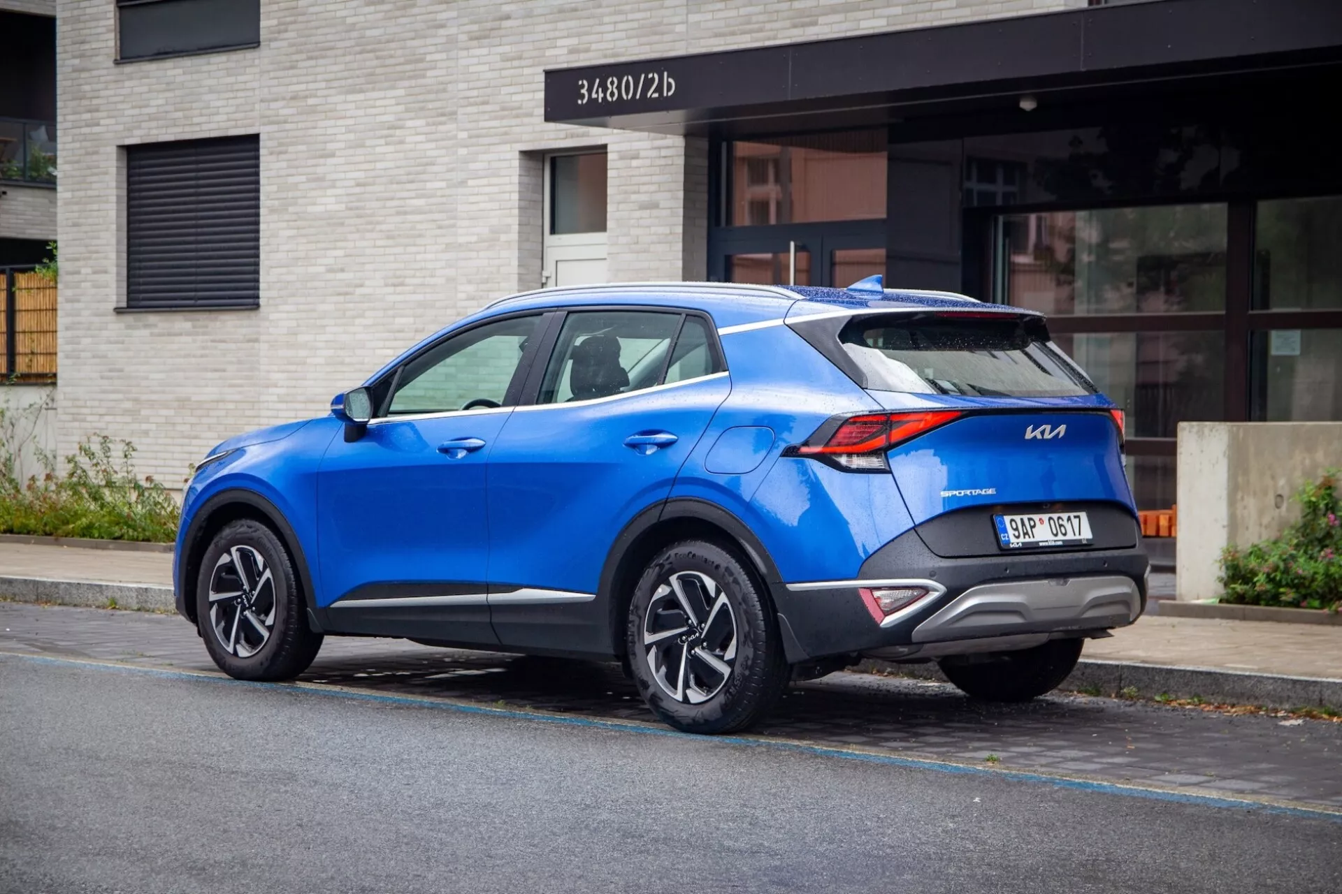 SUV crossover moderno Kia Sportage 2022 estacionado en una calle de la ciudad, mostrando vista trasera de tres cuartos con distintivas luces traseras, alerón trasero deportivo, ruedas de aleación de múltiples radios, color azul vibrante y posibles características tecnológicas modernas.